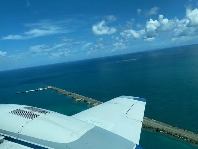 Aerial view from an airplane showing the wing, ocean, and a strip of land with a pier