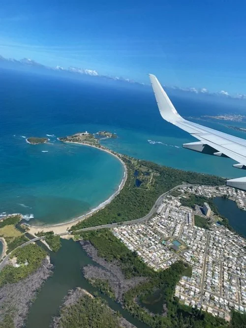 Aerial view from an airplane window showing a coastal area with a beach, blue ocean, green islands, and a nearby town, with the airplane wing visible in the top right corner.