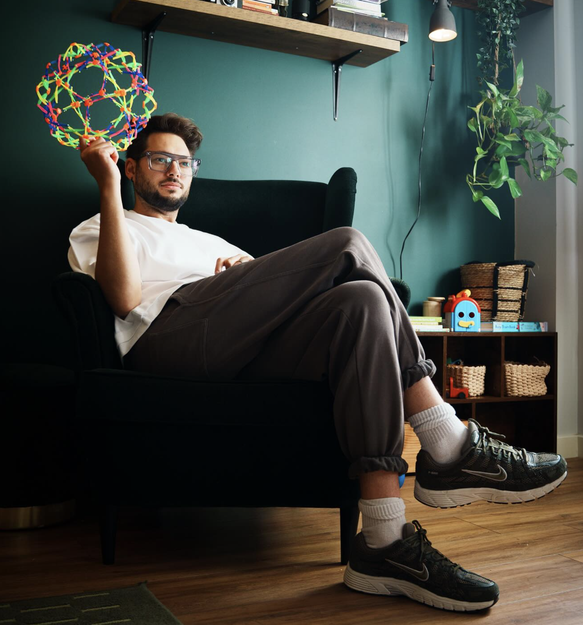 Young man relaxing on a black armchair in a cozy living room, holding a colorful molecular model, with a green wall, a wooden shelf, a potted plant, and children's toys in the background.