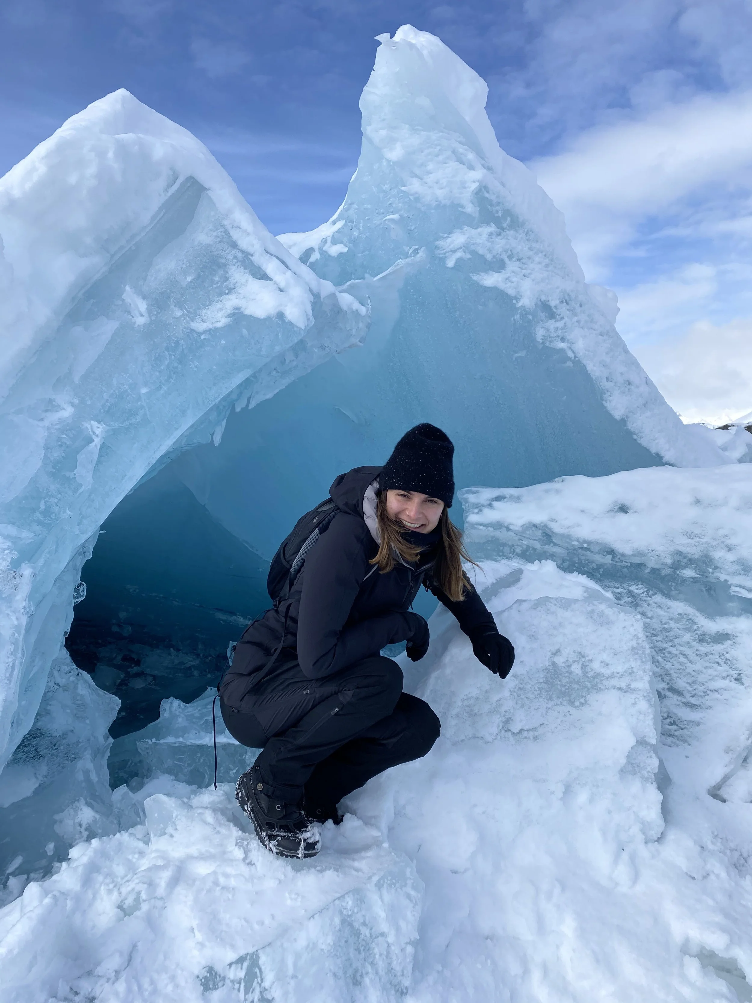 Woman in winter gear kneeling on snow in front of large ice formations on a snowy landscape.