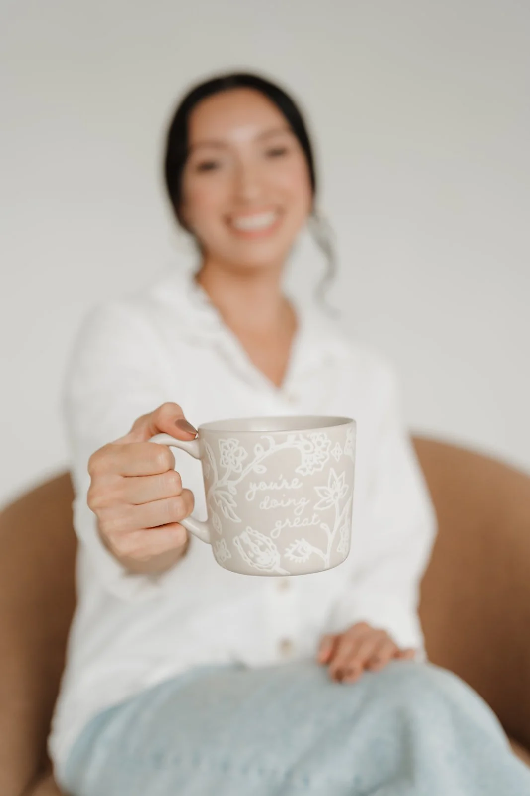 Woman in white shirt sitting on a brown chair, holding out a mug with floral patterns and the words 'you're doing great' written on it, smiling.