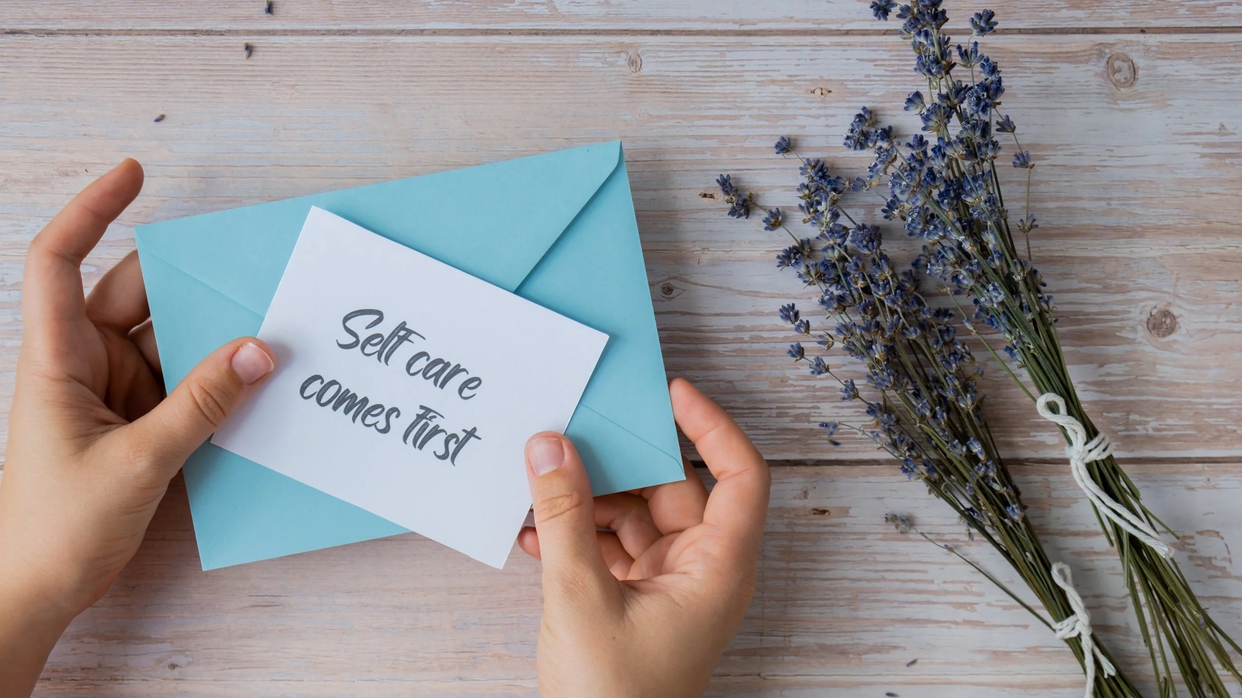 A pair of hands holding an envelope and a note card that says "Self care comes first," with a bunch of lavender flowers on a wooden surface.