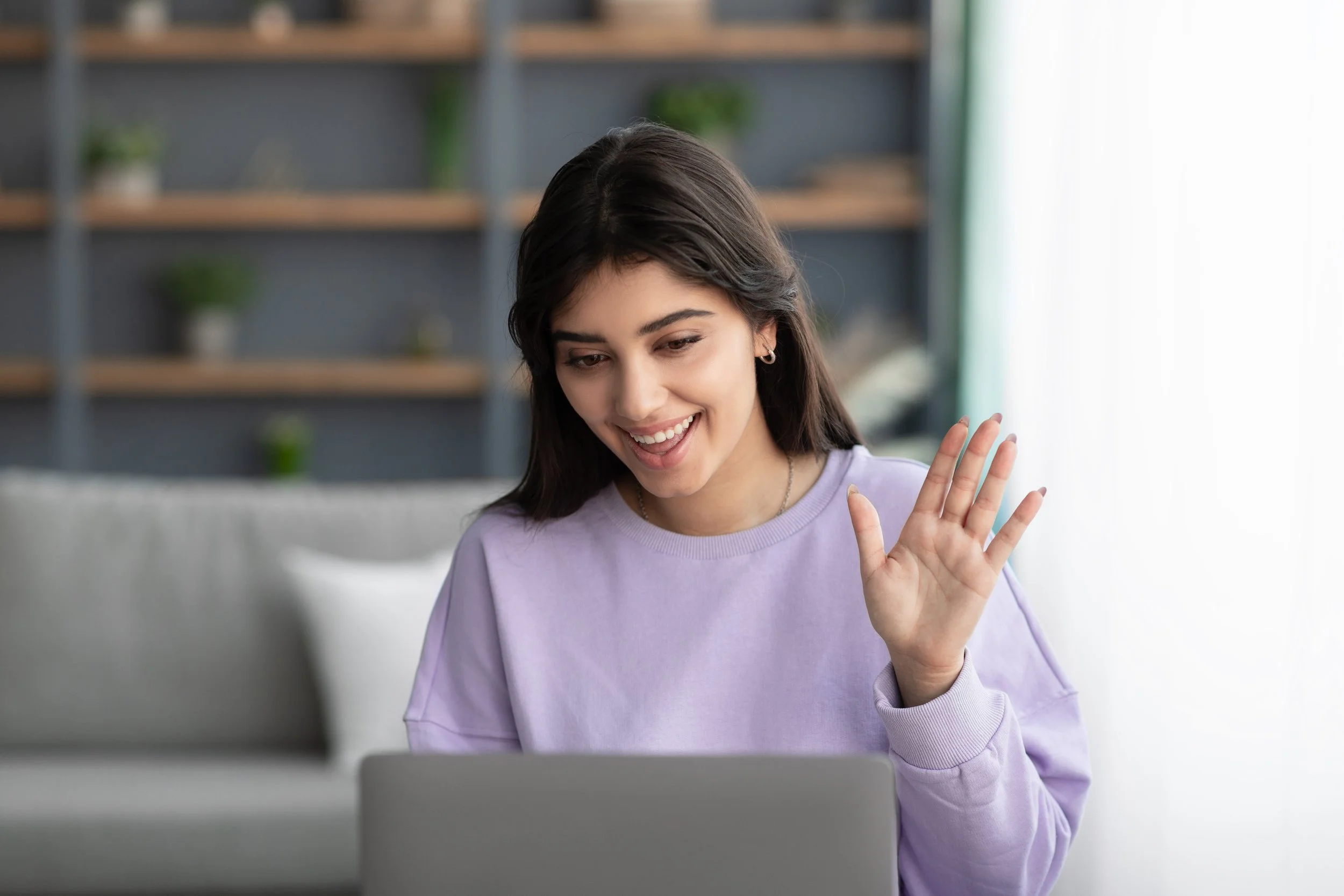 Young woman smiling and waving at a laptop, sitting in a cozy, well-lit room with bookshelves and plants in the background.