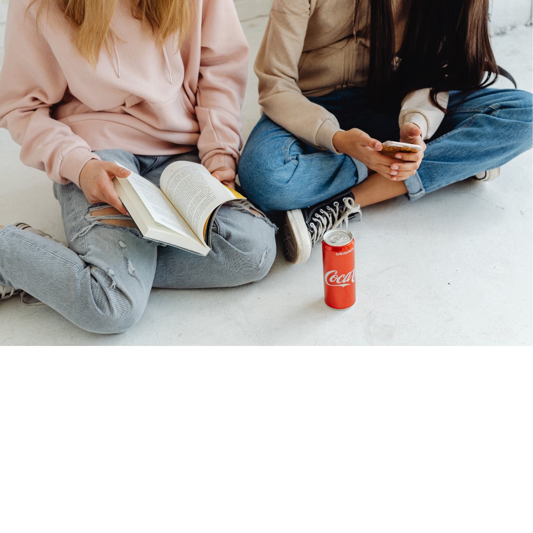 Two people sitting on the floor, one reading a book and the other using a smartphone, with a can of Coca-Cola between them.