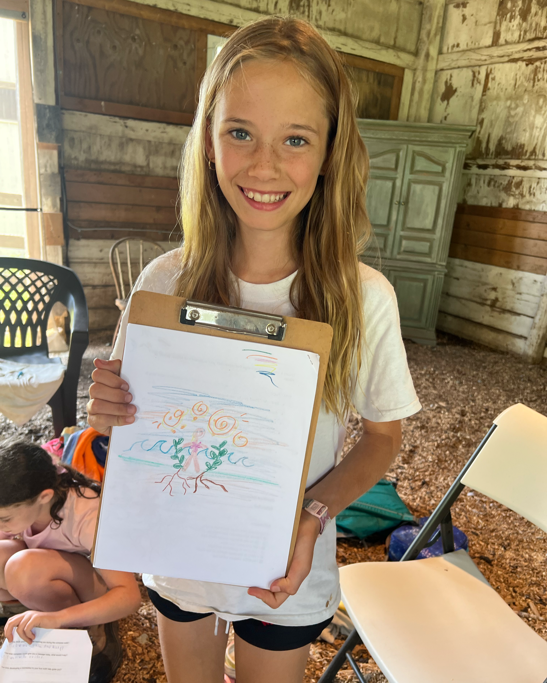 A young girl with long red hair, blue eyes, and freckles is smiling at the camera while holding a clipboard with a colorful drawing that depicts a tree with pink flowers, roots, and blue sky. She is in a rustic wooden room with a green cabinet in the