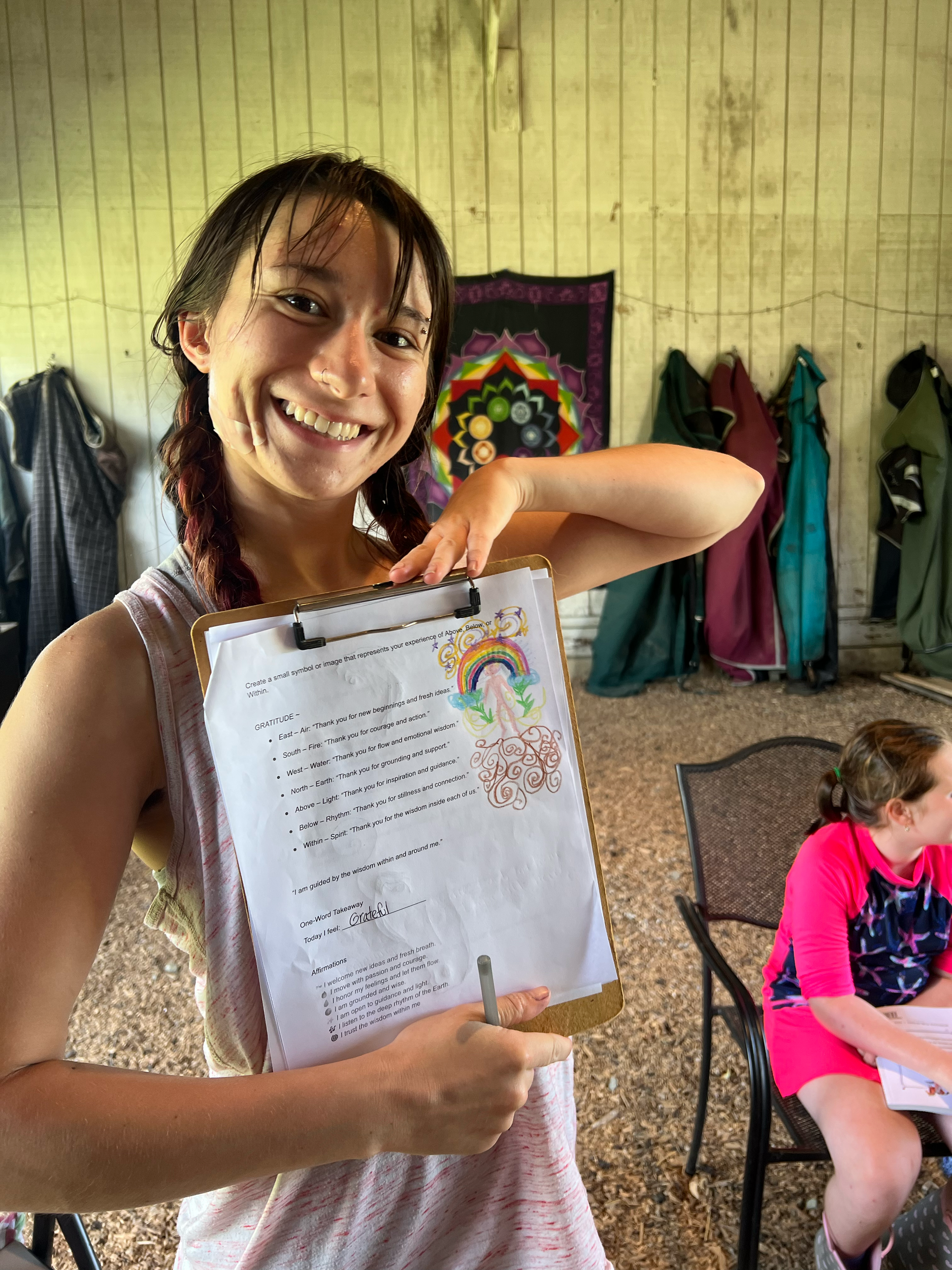 A smiling woman with braided hair holds a clipboard with a colorful drawing of a rainbow and roots, and a sheet of paper with gratitude and affirmations, in a rustic indoor setting with backpacks hanging on the wall.