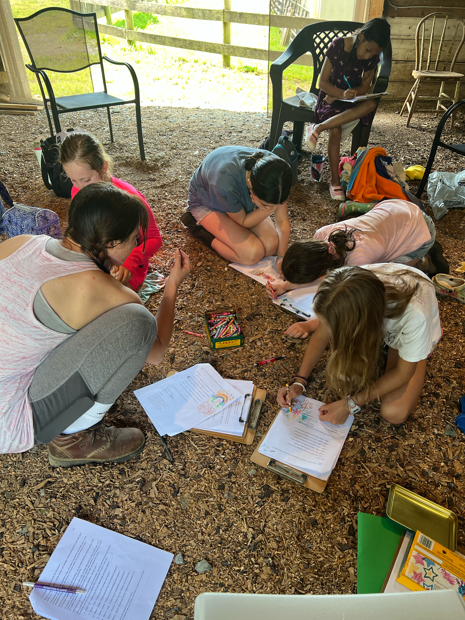 Group of children doing arts and crafts at a booth, with some drawing and coloring on paper, in a rustic setting with chairs and outdoor fencing.