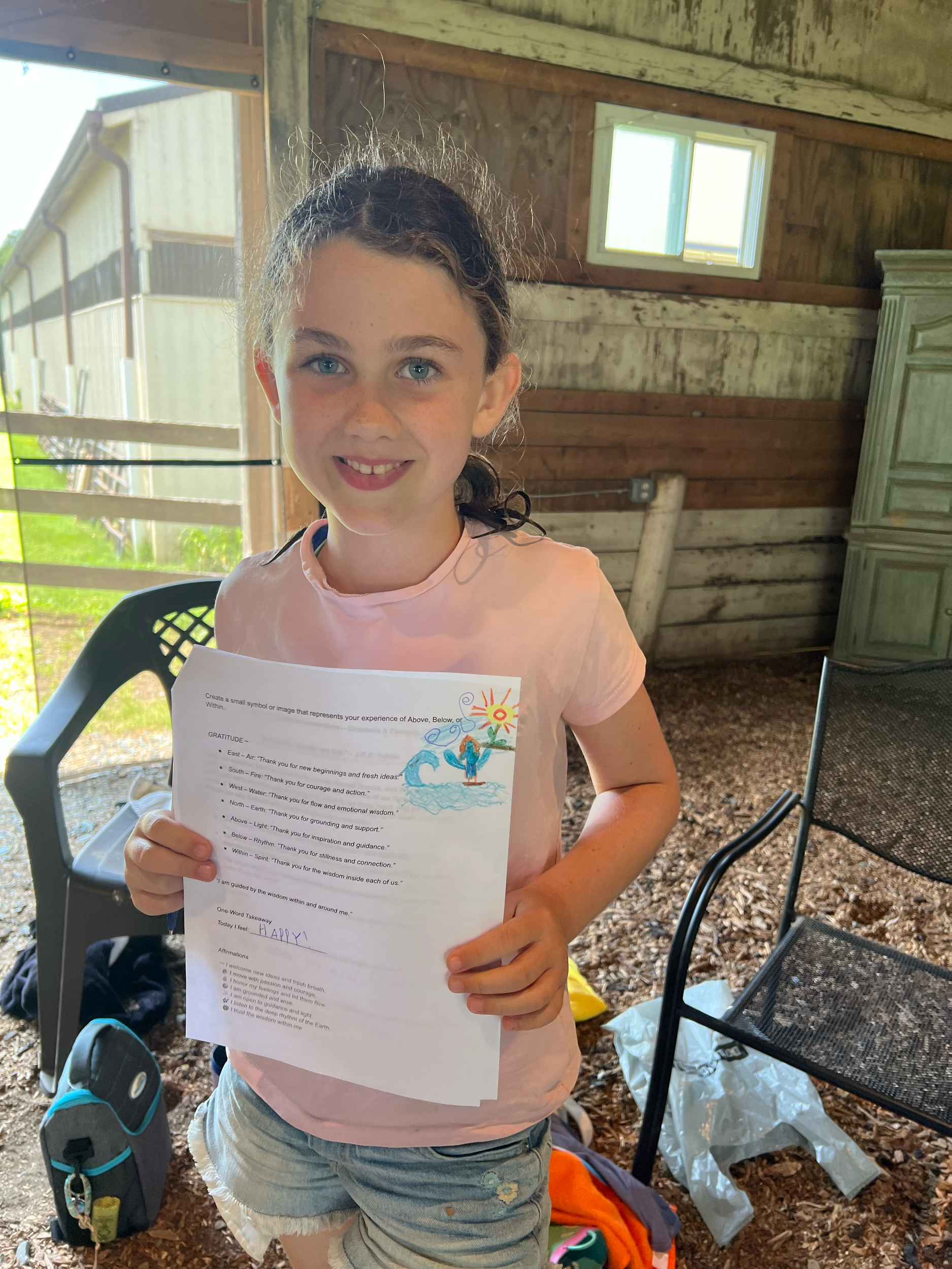 A young girl with curly brown hair and blue eyes stands inside a rustic wooden structure holding a piece of paper with writing and a colorful drawing. She is smiling and wearing a light pink shirt and light-colored shorts. Behind her, there are chair