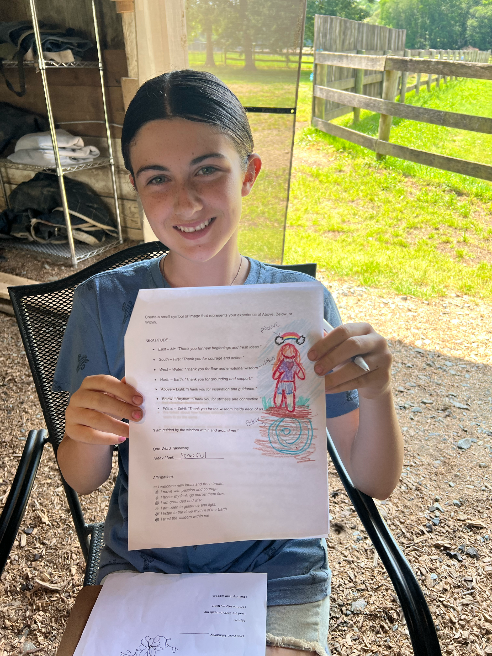 A young girl with dark hair in a wet style, sitting outdoors in a shaded area, smiling and holding a piece of paper with a colorful drawing of a girl with red hair, a rainbow headband, and the words 'Above' and 'Below' written on it.