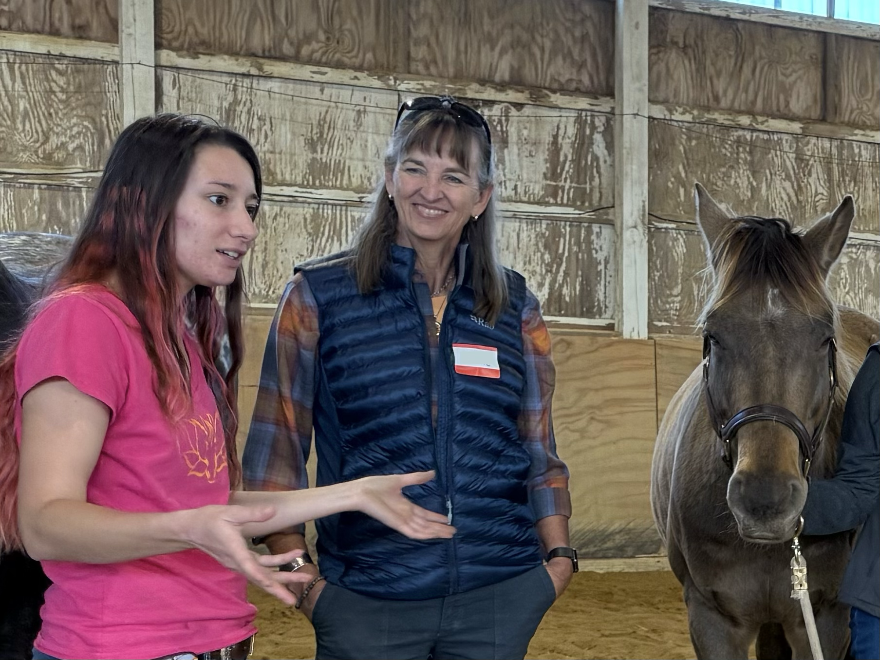 Two women and a horse inside a wooden stable. The young woman on the left, wearing a pink shirt, is talking, while the older woman in a blue vest listens. The horse is on the right, facing forward with a harness.