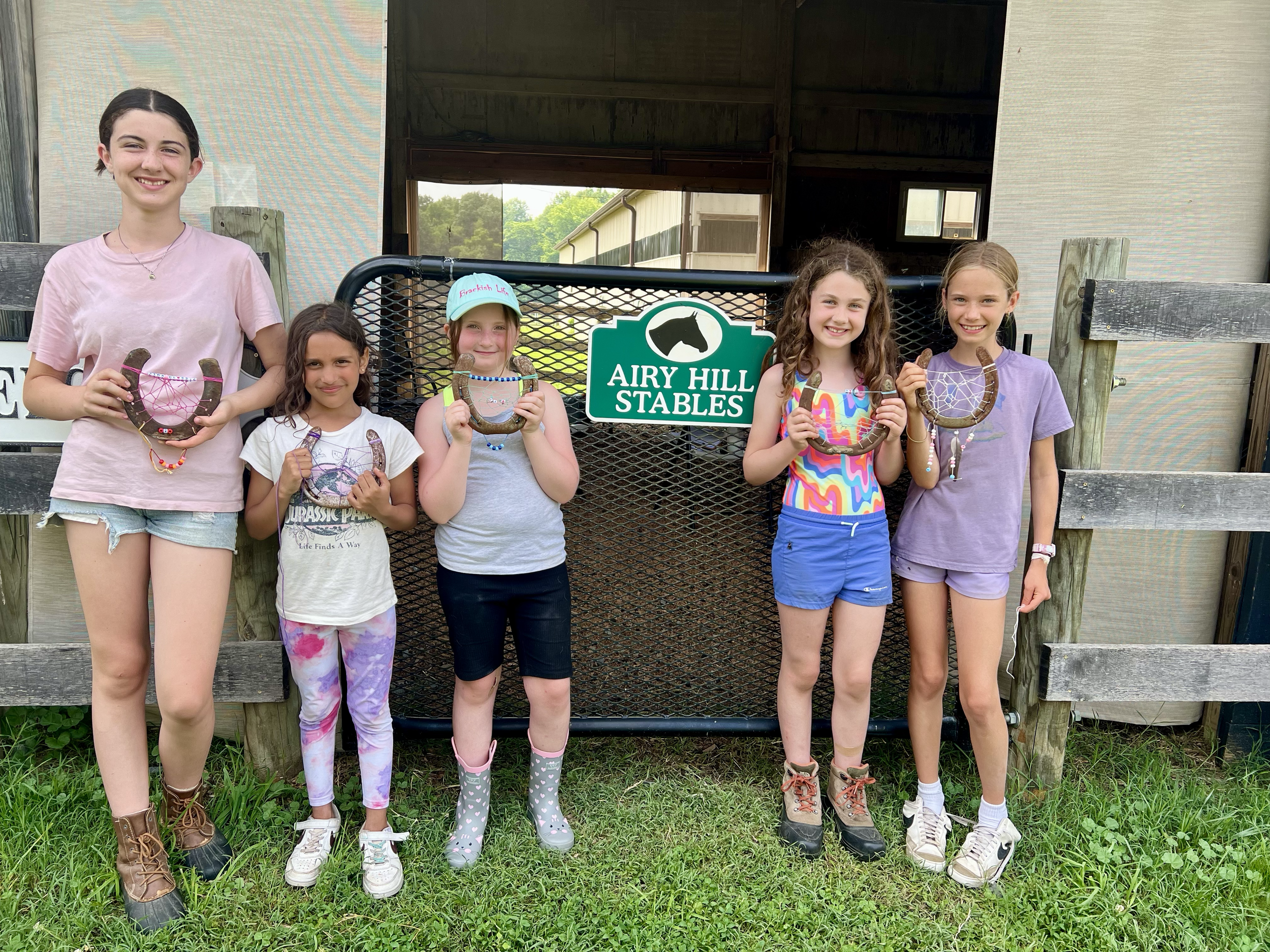 A group of six young girls standing outdoors in front of a sign that reads "Airy Hill Stables". They are holding horseshoes decorated with beads and strings, dressed casually in colorful clothes, and smiling.