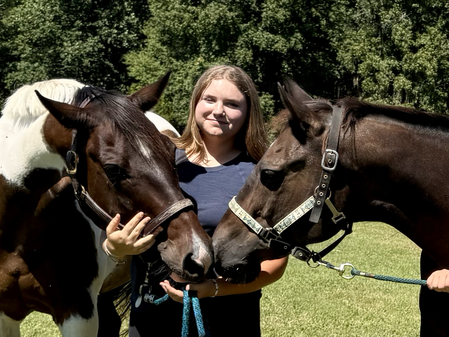 A young woman standing outdoors with two horses, holding their bridles, on a sunny day with trees in the background.