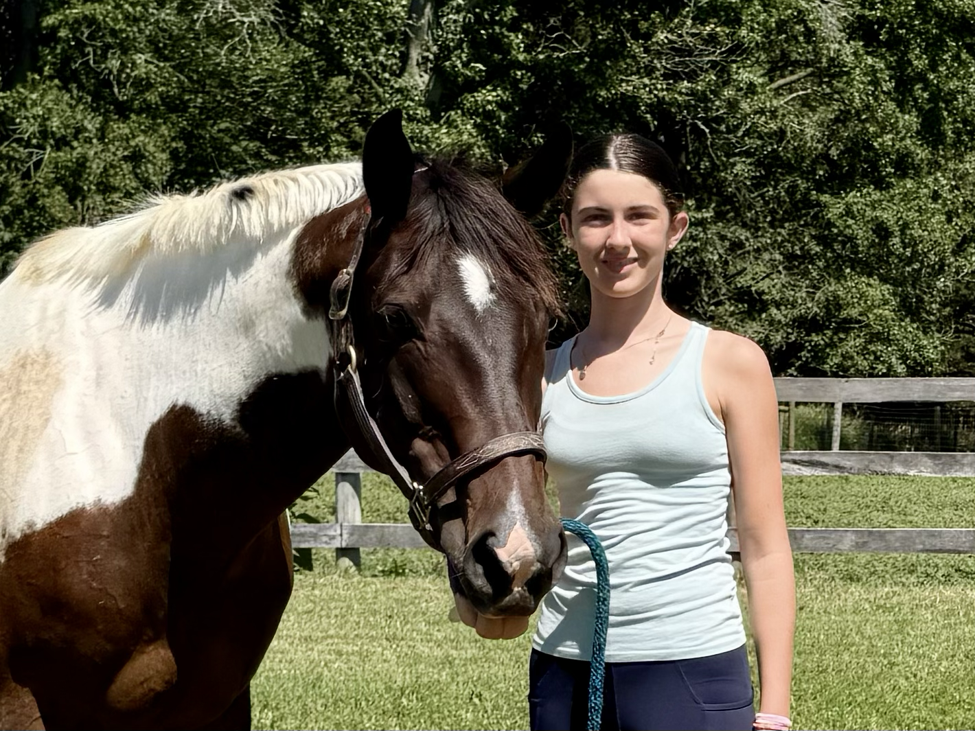A young woman standing outdoors in a grassy area with a chestnut horse with a white star on its forehead. She is smiling and holding the horse's lead rope, with a wooden fence and green trees in the background.