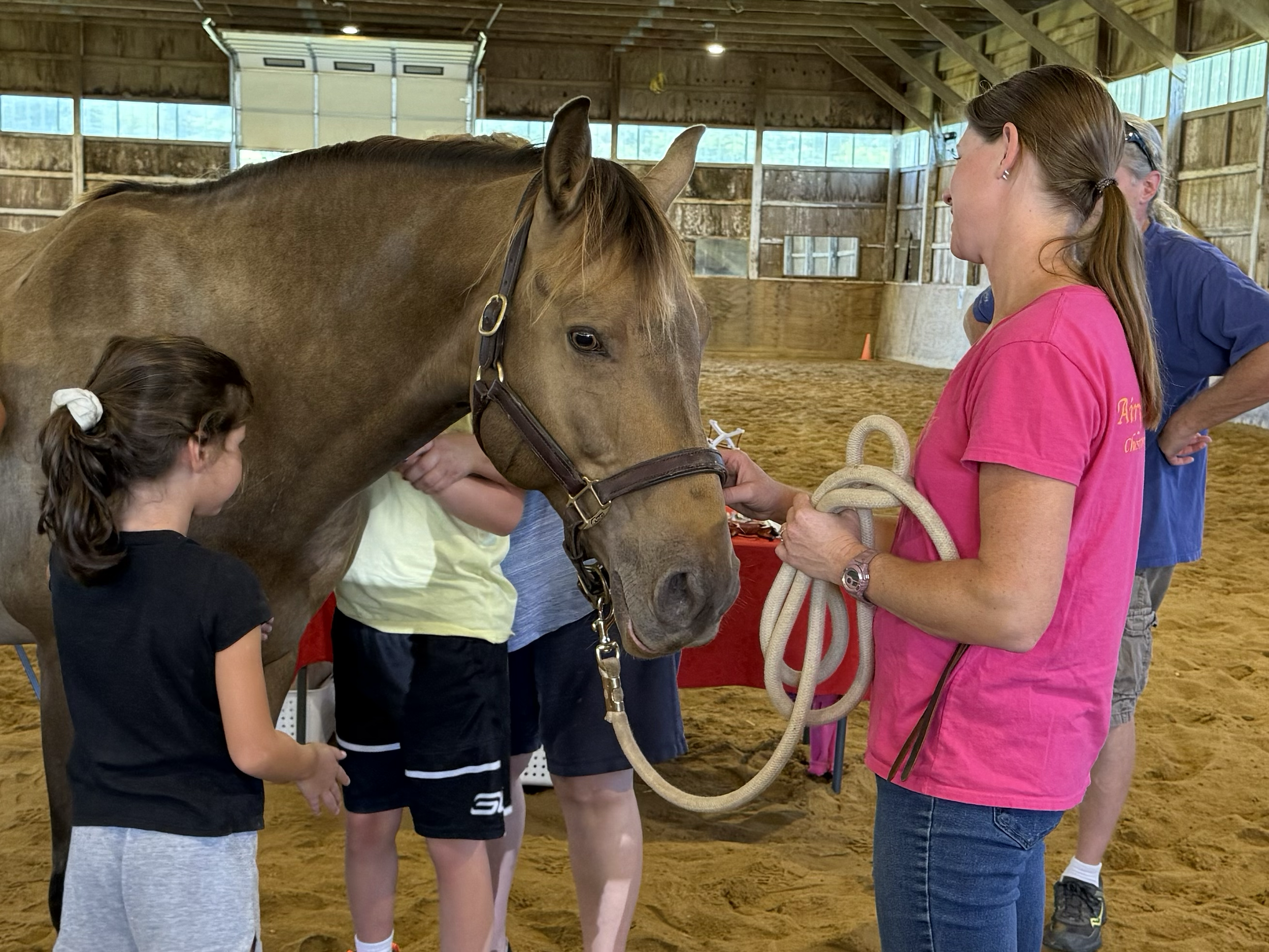 A woman in a pink shirt holding a lead rope while a group of children examine a tan horse in an indoor riding arena.