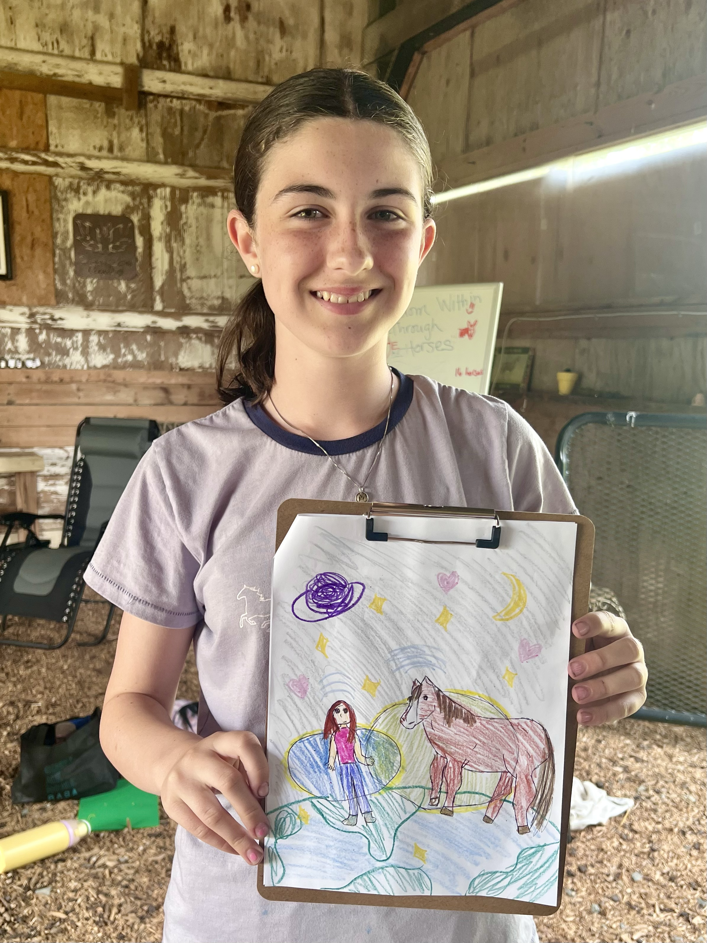 A young girl with brown hair in a ponytail, smiling and holding a clipboard with a colorful drawing of a girl and a horse, with stars, hearts, moons, clouds, and a purple swirl in the sky.
