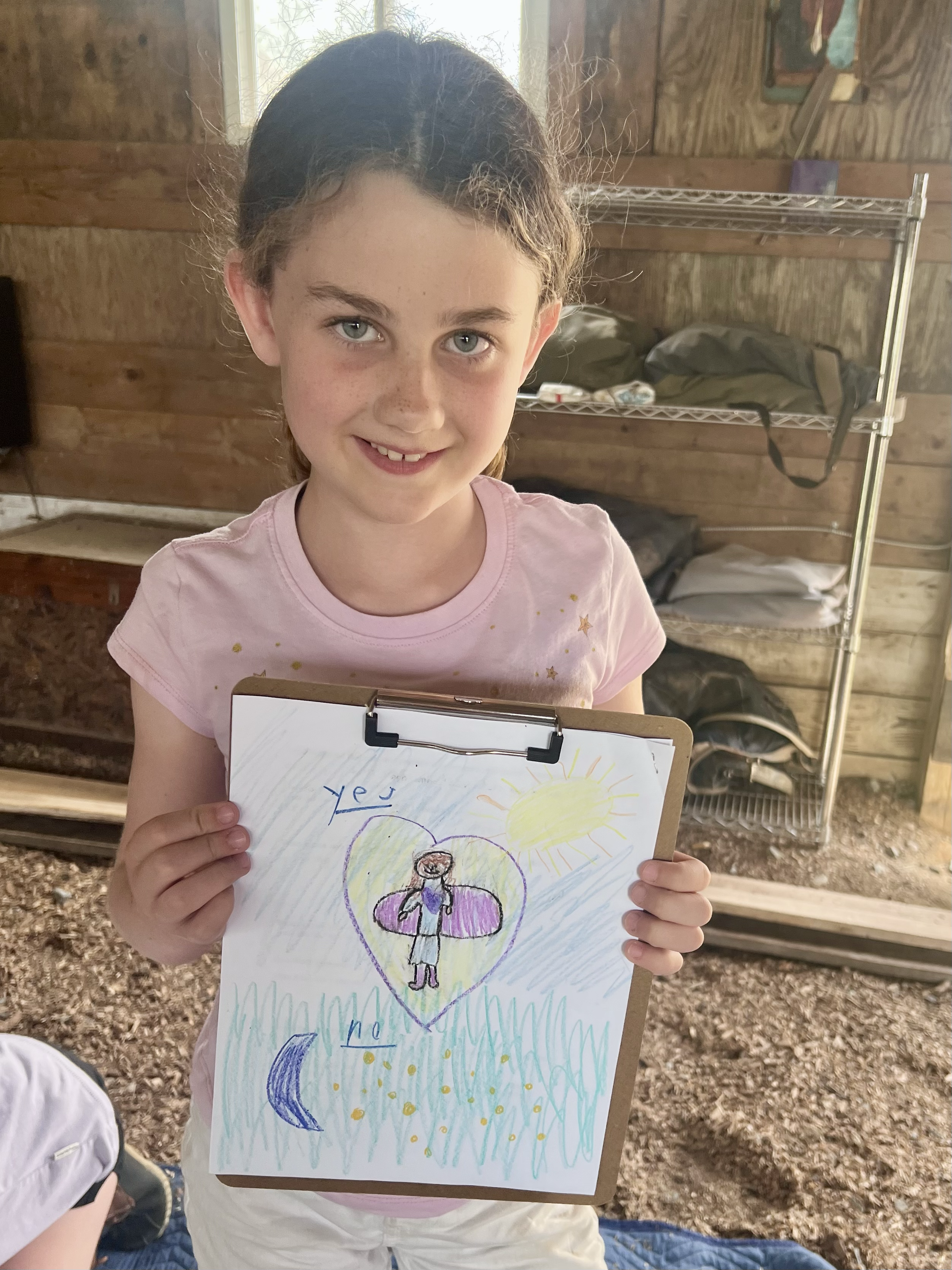 A young girl with light skin, curly brown hair, and blue eyes holding a drawing on a clipboard, smiling at the camera inside a rustic wooden room with a metal shelf and outdoor scenery visible through a window.