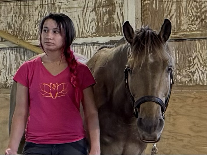 A girl with long dark hair in a pink shirt standing next to a brown horse inside a wooden stable.