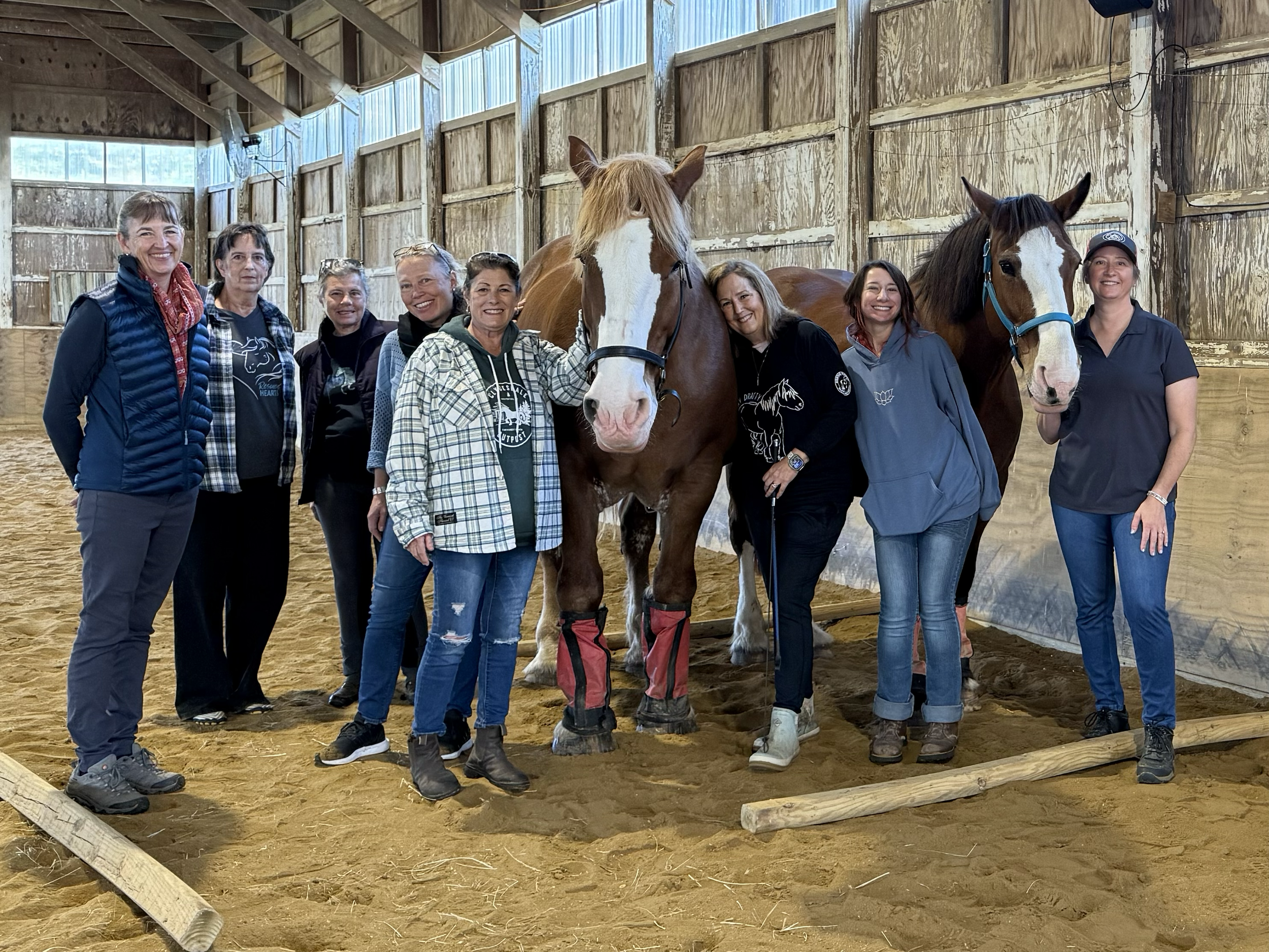 Group of women standing in a riding arena with two horses, smiling for a photo.