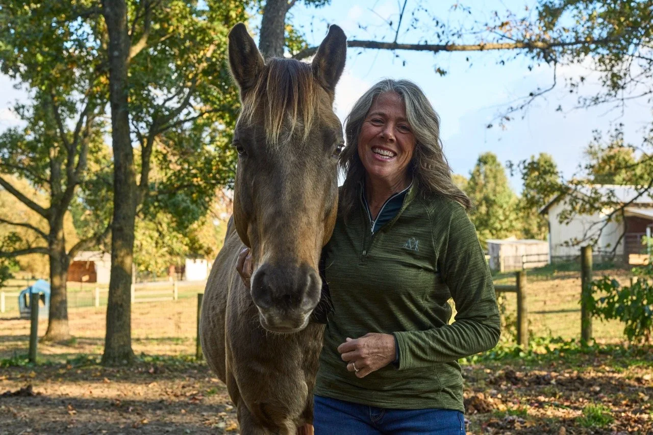 A woman smiling and standing next to a brown horse outdoors in a park-like setting with trees and a fence.