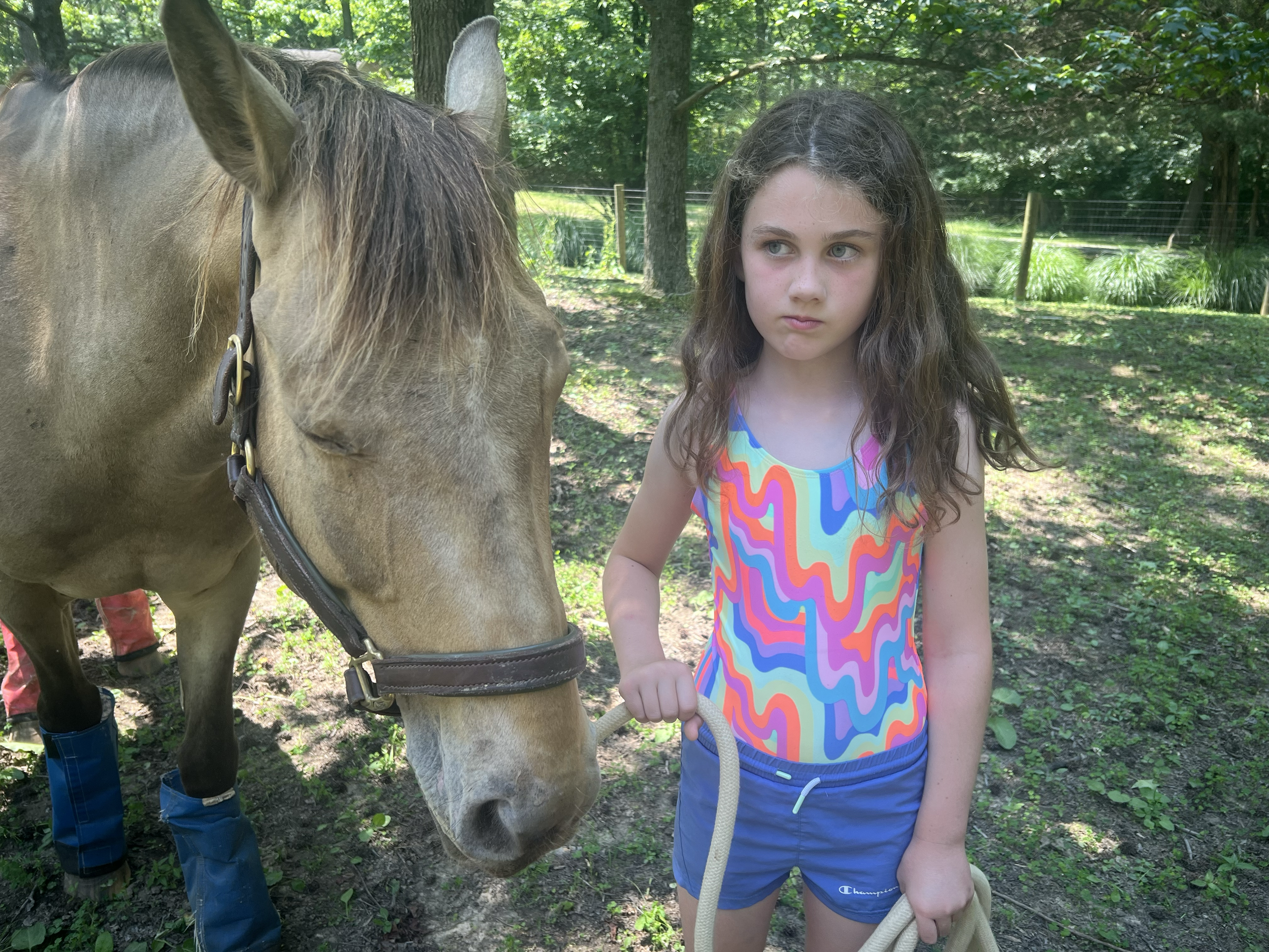 A young girl with long curly brown hair, wearing a colorful print tank top and blue shorts, is holding a rope and standing next to a light brown horse with a dark mane in a wooded outdoor area.