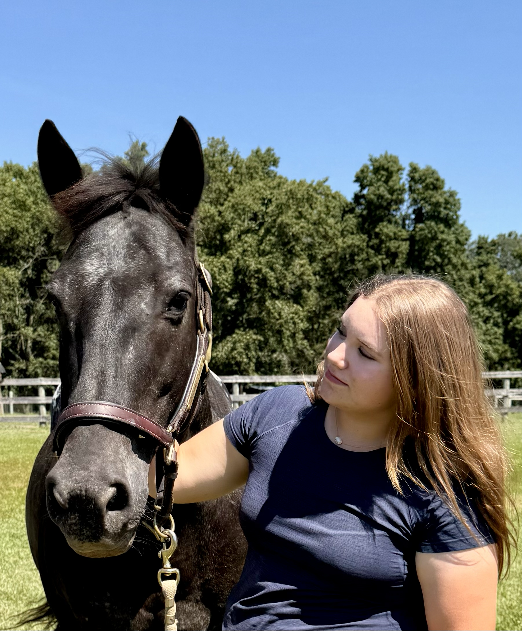 A woman with long red hair in a navy blue shirt standing next to a black horse with a white star on its forehead in an outdoor grassy area with a fence and trees in the background.