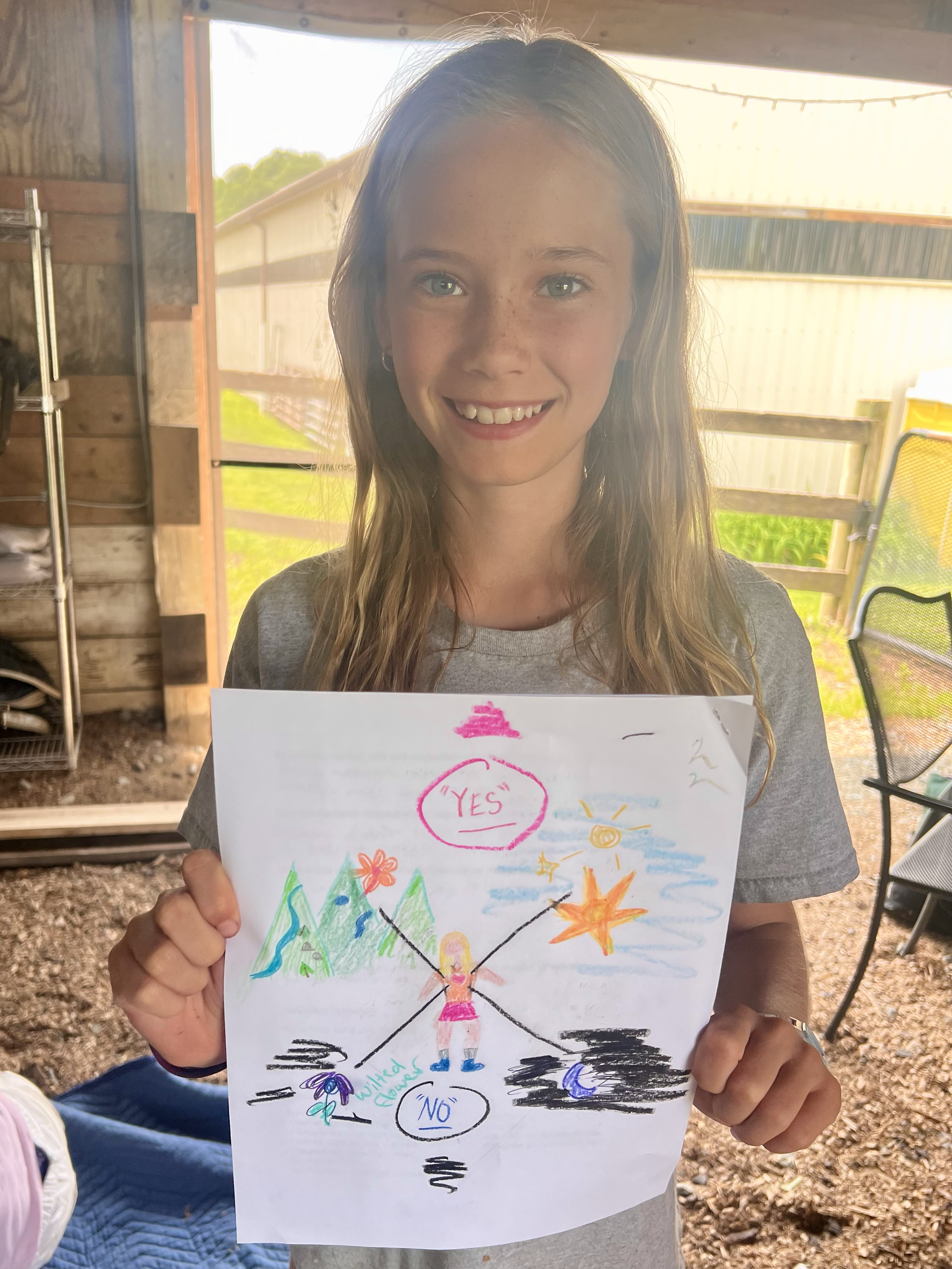 A girl with long, light brown hair smiling and holding a colorful drawing. The drawing features a central figure with arms raised, colorful mountains, clouds, stars, and the words 'YES' and 'NO' written in circles, along with other decorative element
