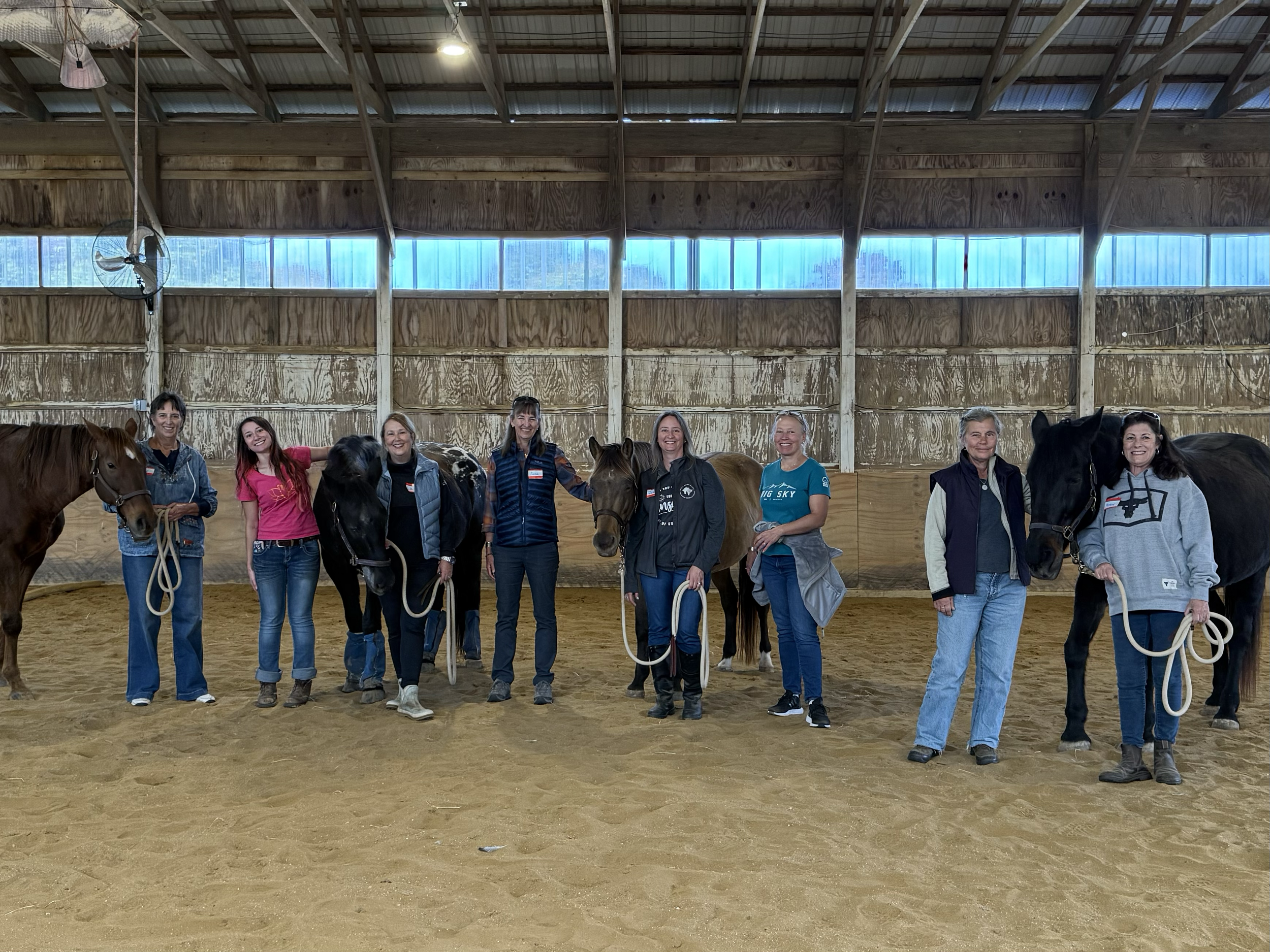 Group of women standing with horses inside an indoor riding arena, some holding the horses' reins, smiling at the camera.