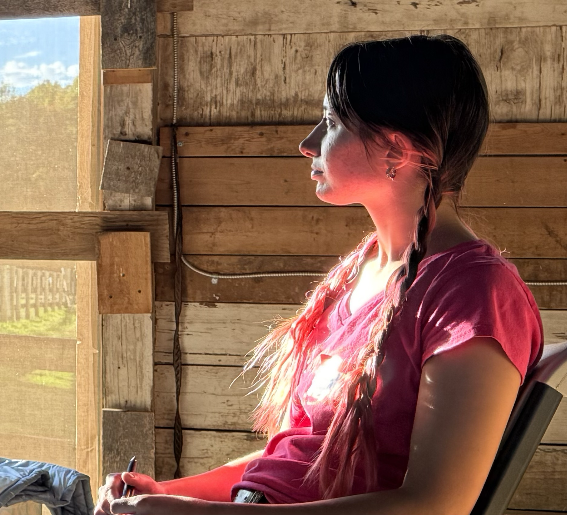 A woman with long braided hair sitting inside a wooden cabin near a window, with sunlight illuminating her face and shoulder.