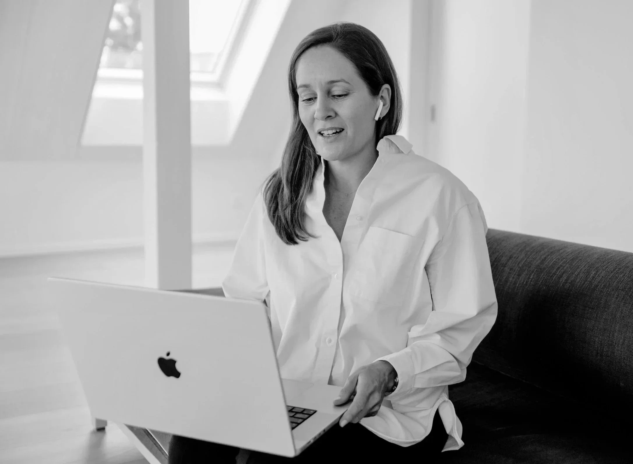 A woman with long hair wearing a white shirt, sitting on a couch, looking at a laptop, with earphones in her ears.