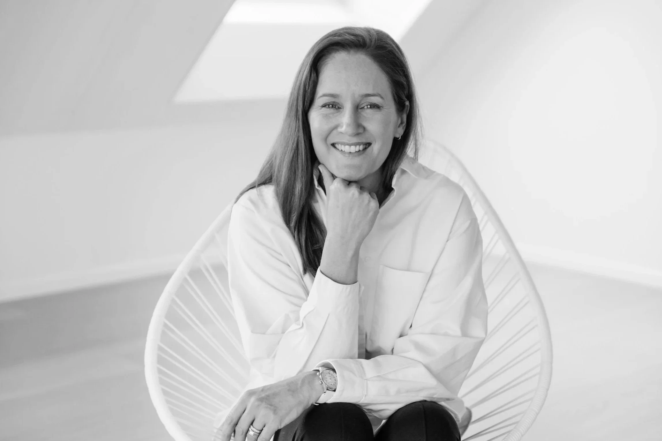 A woman with long hair smiling and sitting in a modern chair, wearing a white shirt and a watch.