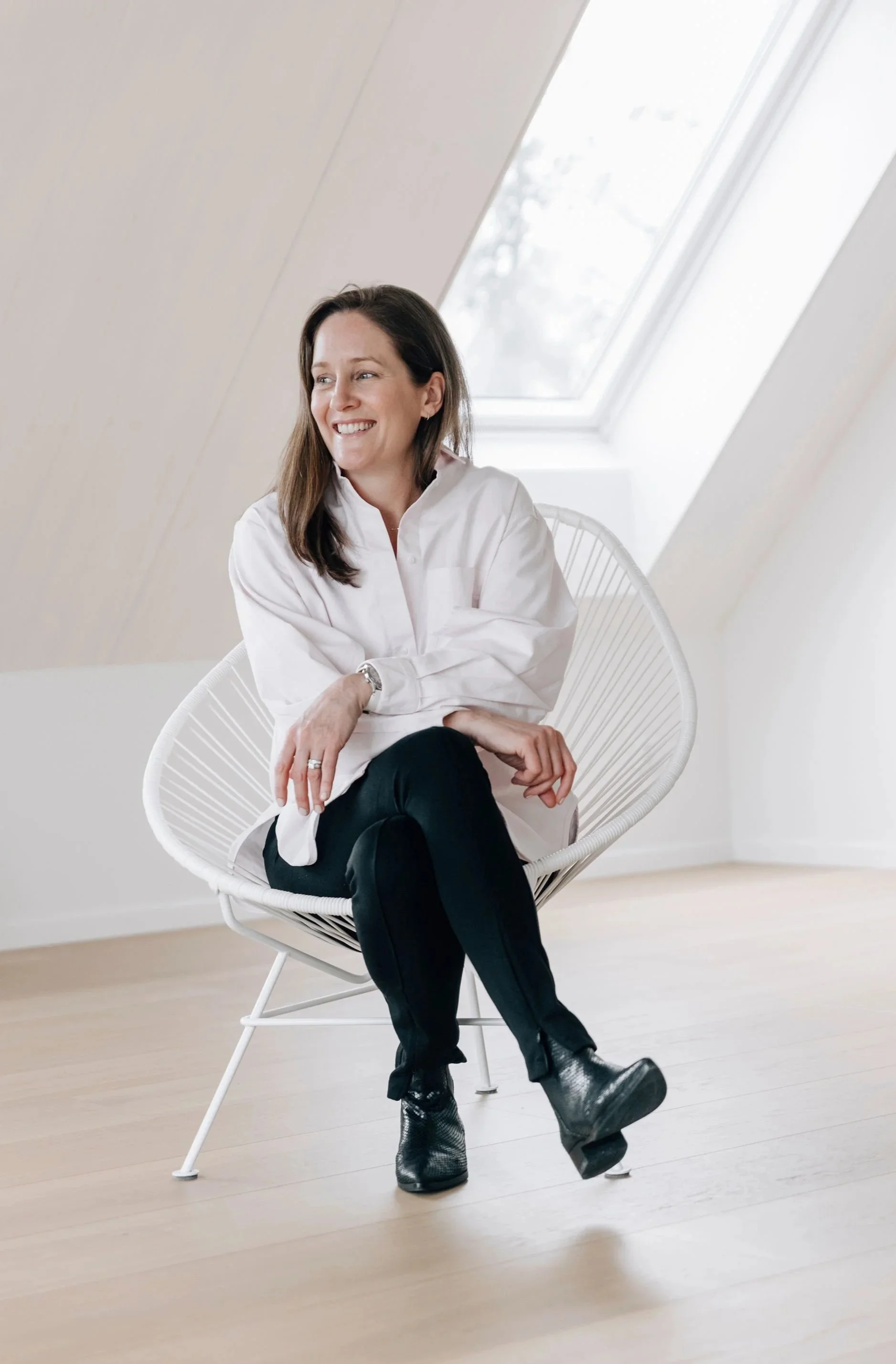 A woman with dark hair, smiling, sitting on a white modern chair in a bright room with a sloped ceiling and a skylight window.