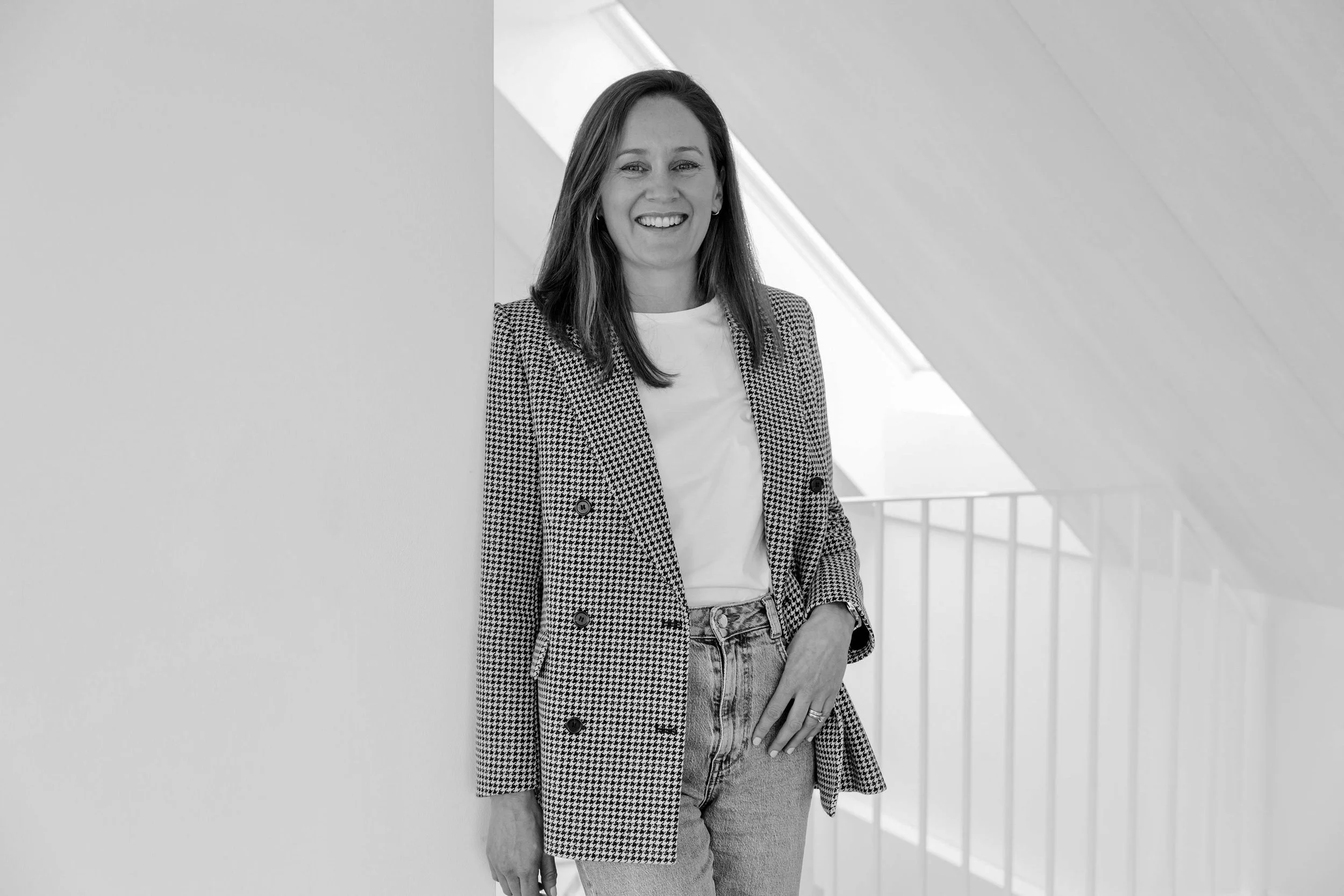 A woman with long hair, smiling, wearing a checkered blazer and jeans, standing near a wall with a modern architectural background.