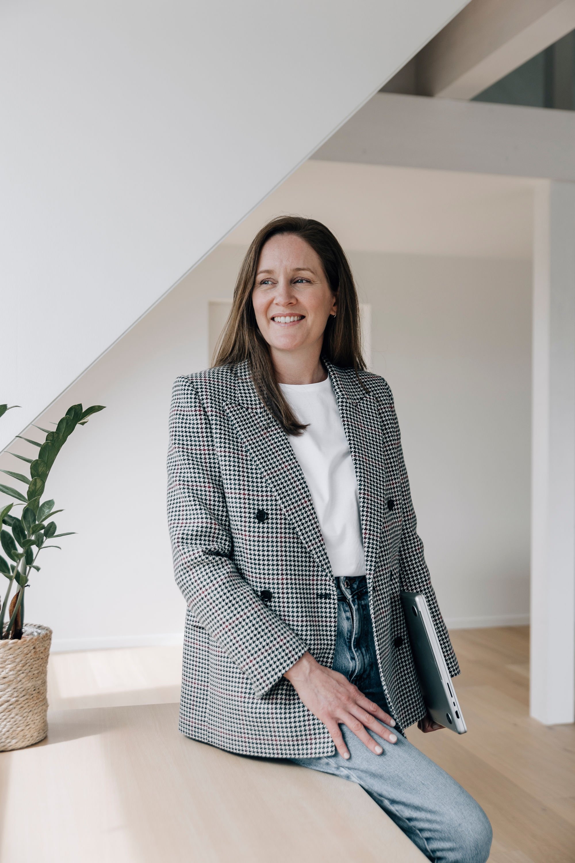 A woman with long brown hair, wearing a checkered blazer and jeans, sitting on a wooden surface holding a closed laptop, smiling in a modern interior with white walls and a potted plant.