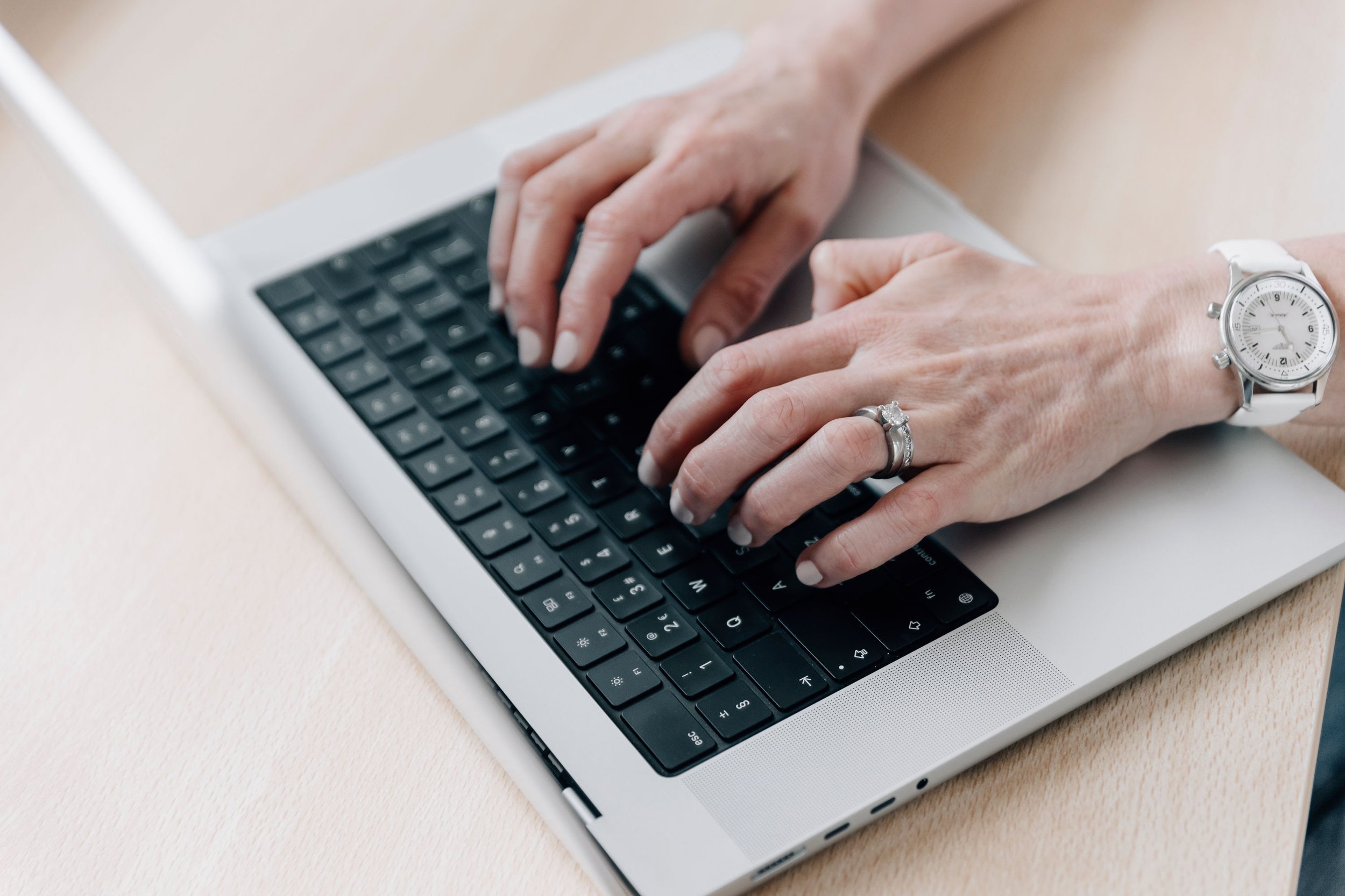 A person typing on a black keyboard of a silver laptop, wearing a white watch and a ring on their left hand.