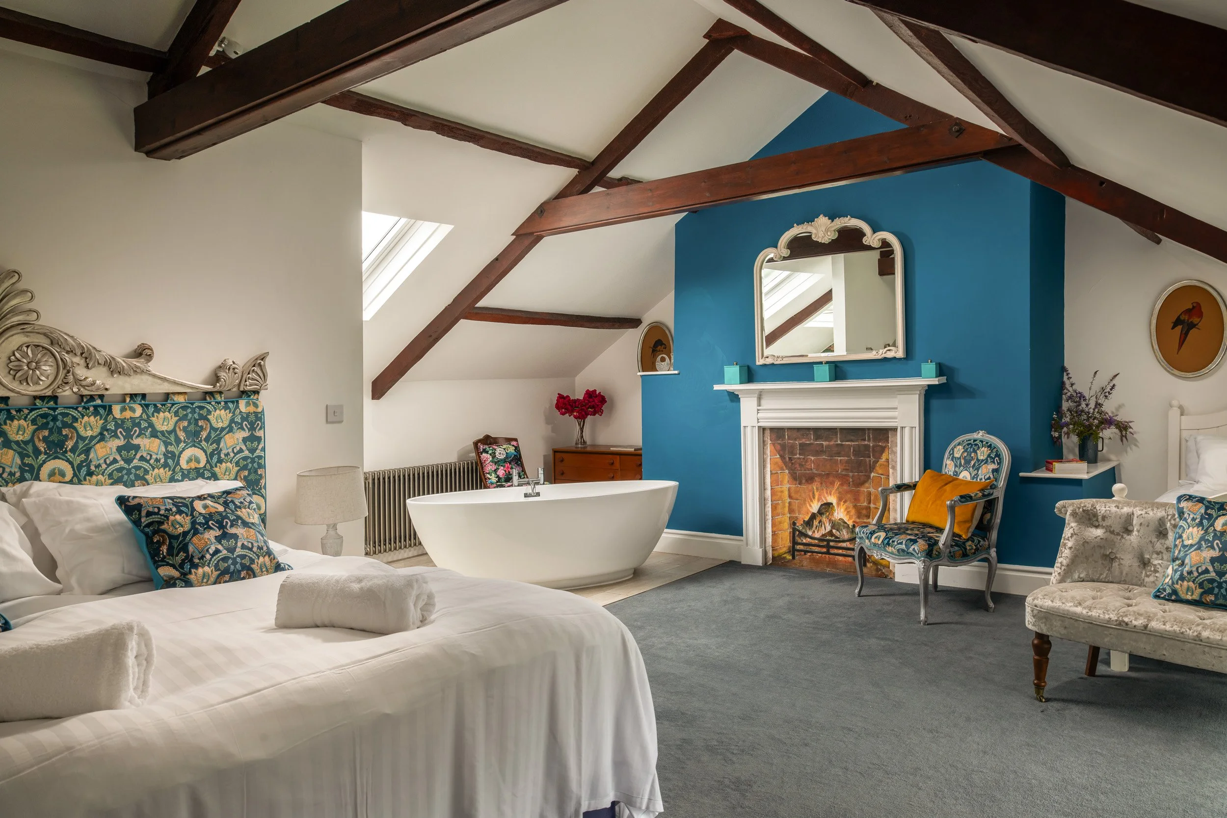 A cozy attic bedroom with a large bed on the left, featuring blue and gold patterned pillows and white linens. A bathtub is positioned near a small chest of drawers, with a floral blanket draped over a chair. The room has a bright fireplace with a mi