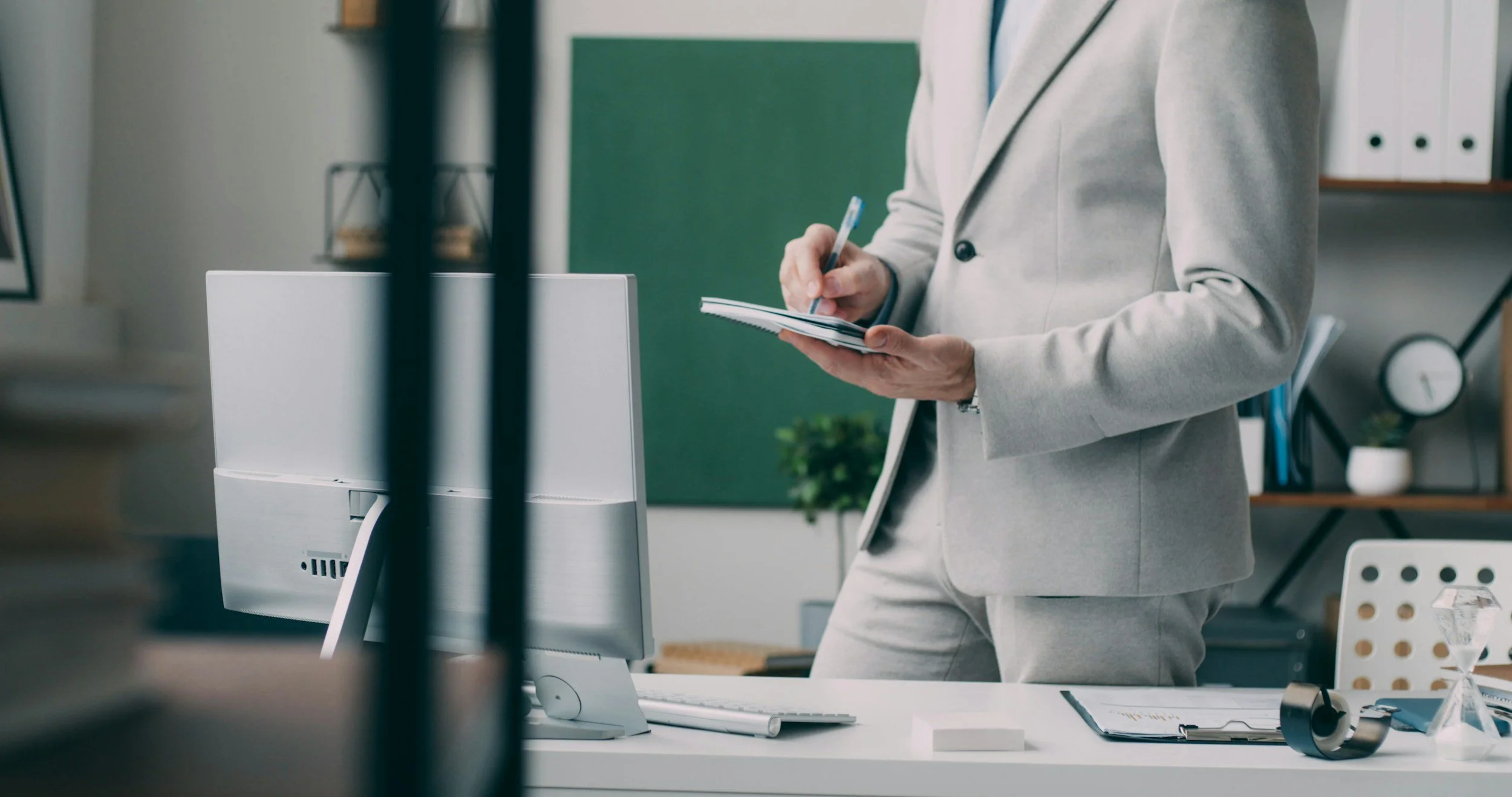 Person in a beige suit standing at a desk writing in a notepad, visible community office or workspace environment.