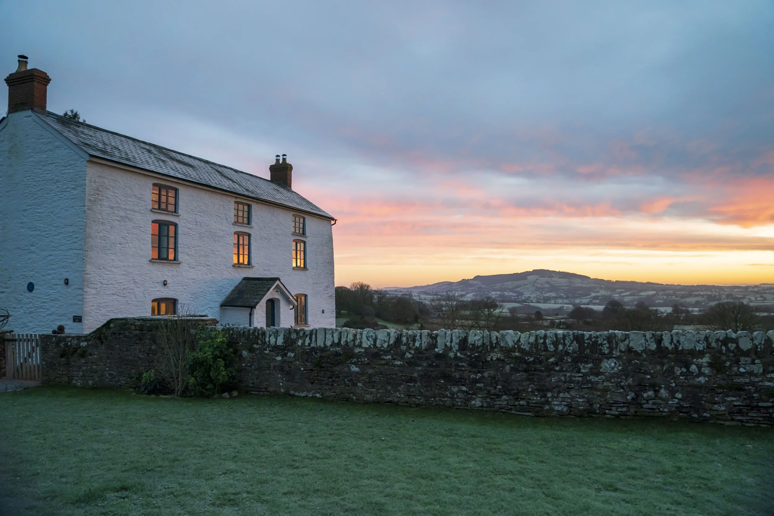 A white house with multiple windows, some illuminated from inside, sitting on a grassy lawn with a stone wall in front. The background features a scenic sunset with colorful clouds and rolling hills in the distance.