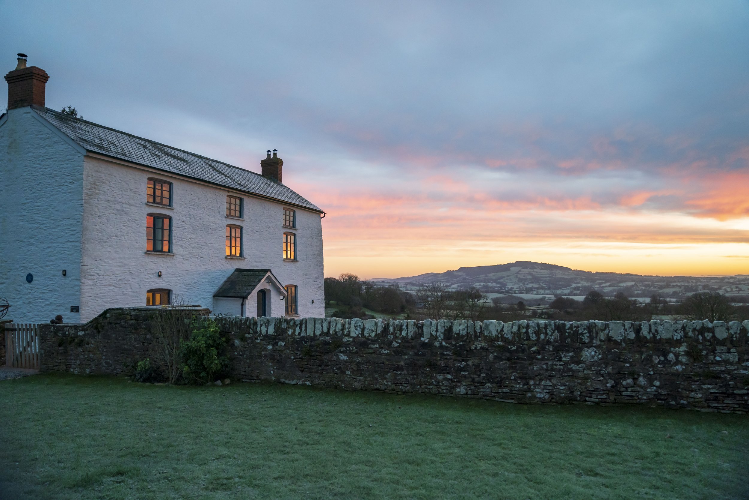 A white house with lit windows at sunset, surrounded by a green lawn and stone wall, with rolling hills and colorful sky in the background.