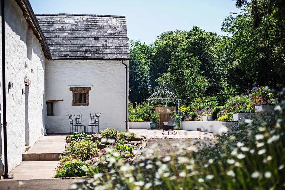 A peaceful garden with a white cottage wall, an outdoor seating area, a decorative birdcage, and lush green trees in the background.