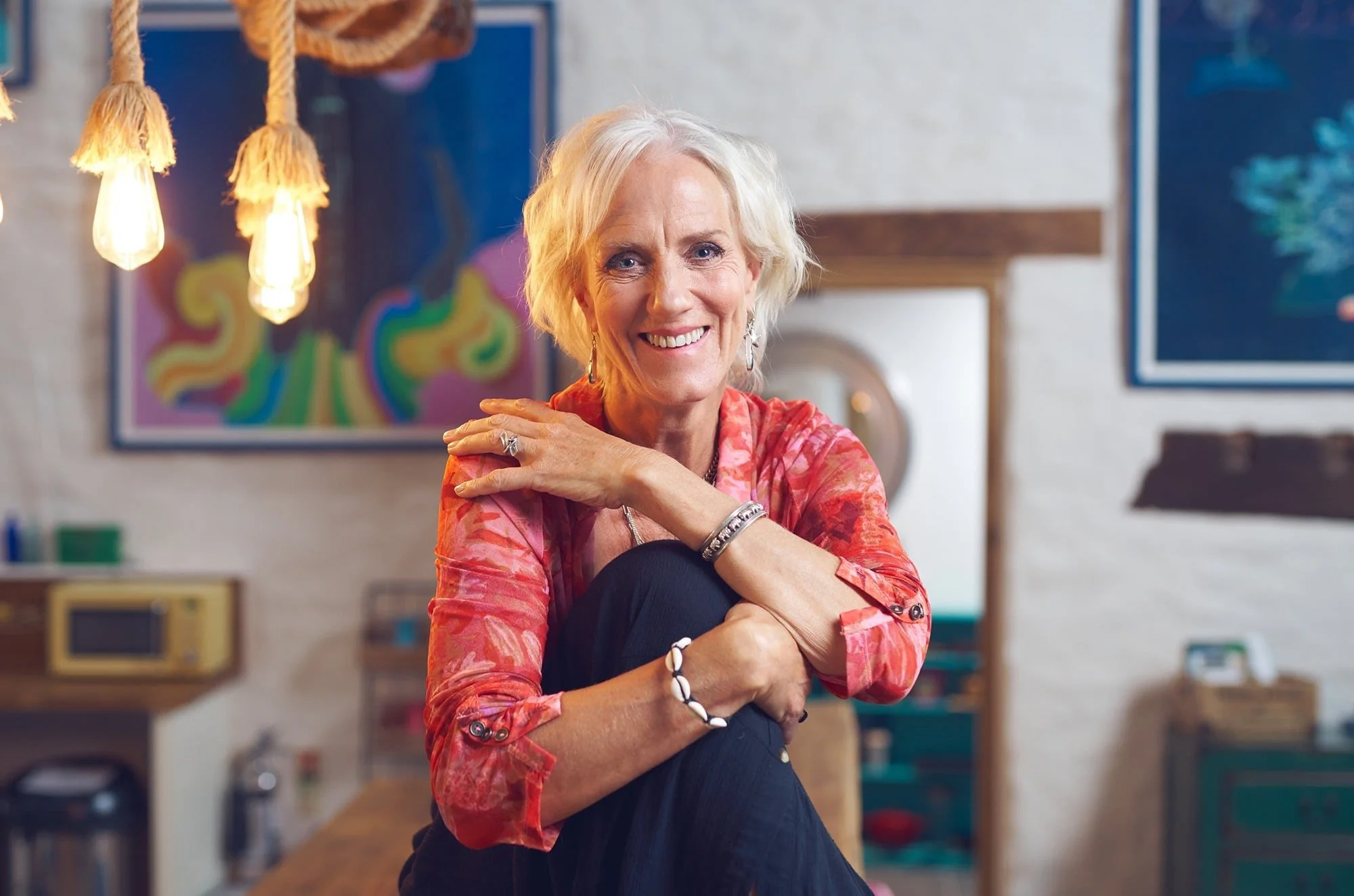 A smiling woman with short blonde hair wearing a red patterned blouse and black pants, sitting in a colorful, artsy room with paintings and hanging lights.