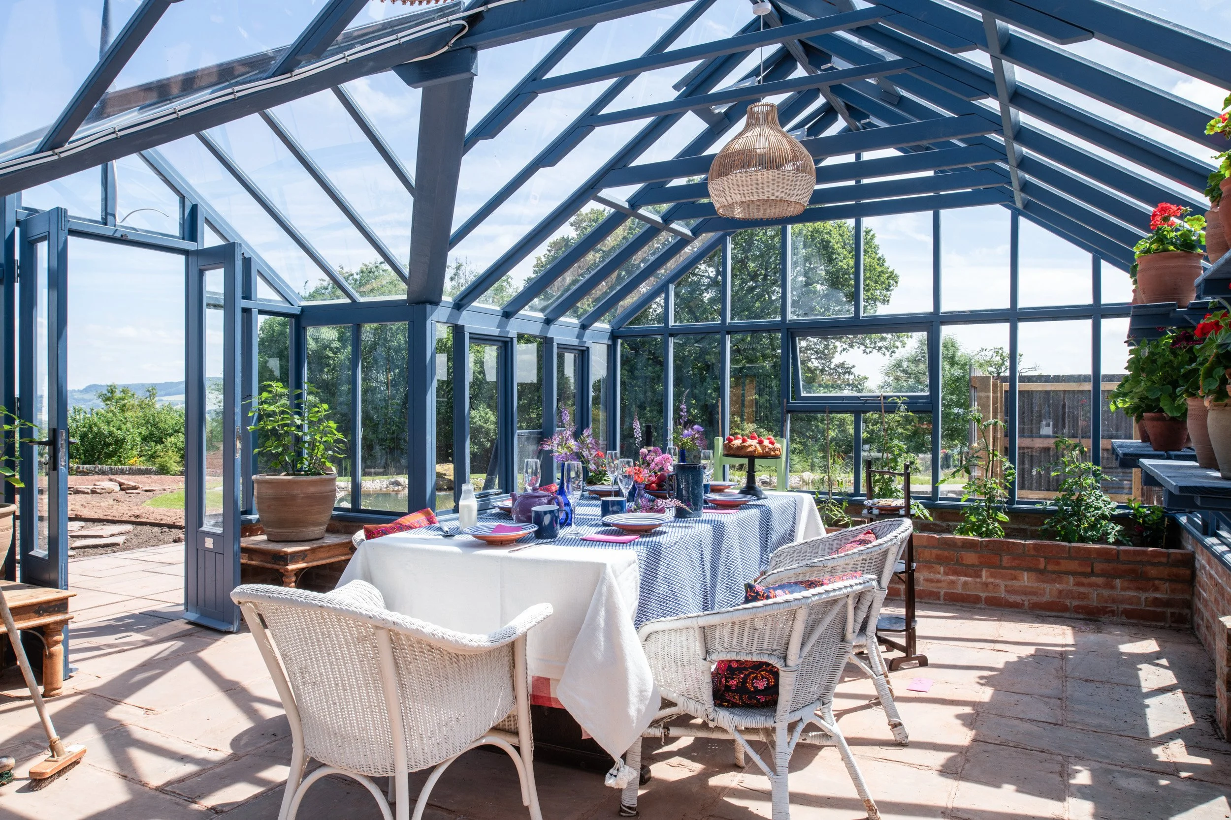 A sunny greenhouse with a table set for a meal, surrounded by potted plants and flowers, with a view of the outdoors through large glass windows.