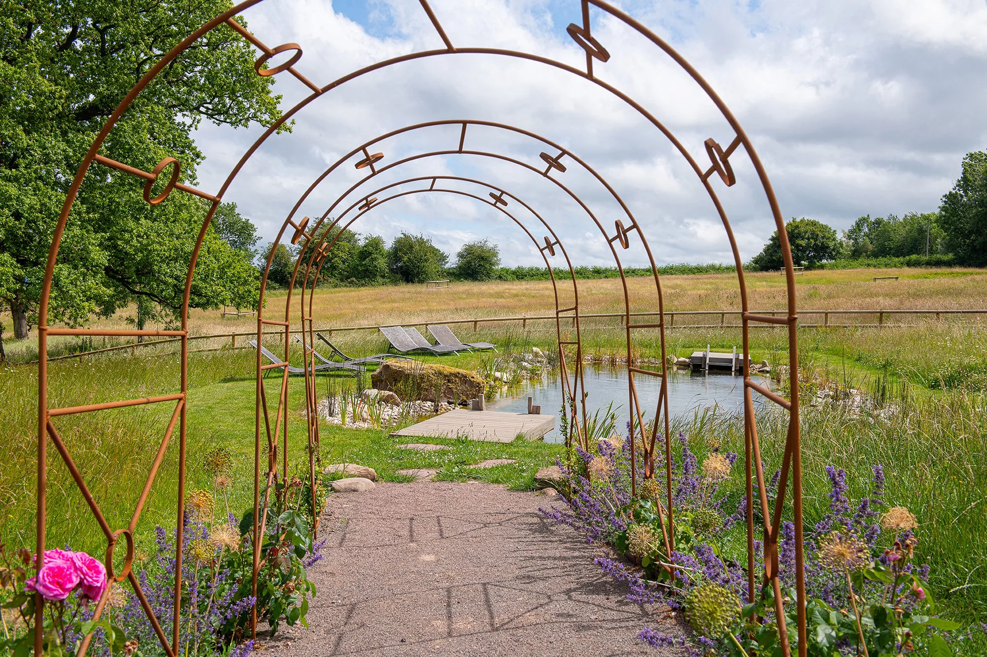 Garden path with brown metal arches overgrown with purple and pink flowers, leading to a small pond with lounge chairs and lush green fields under partly cloudy sky.