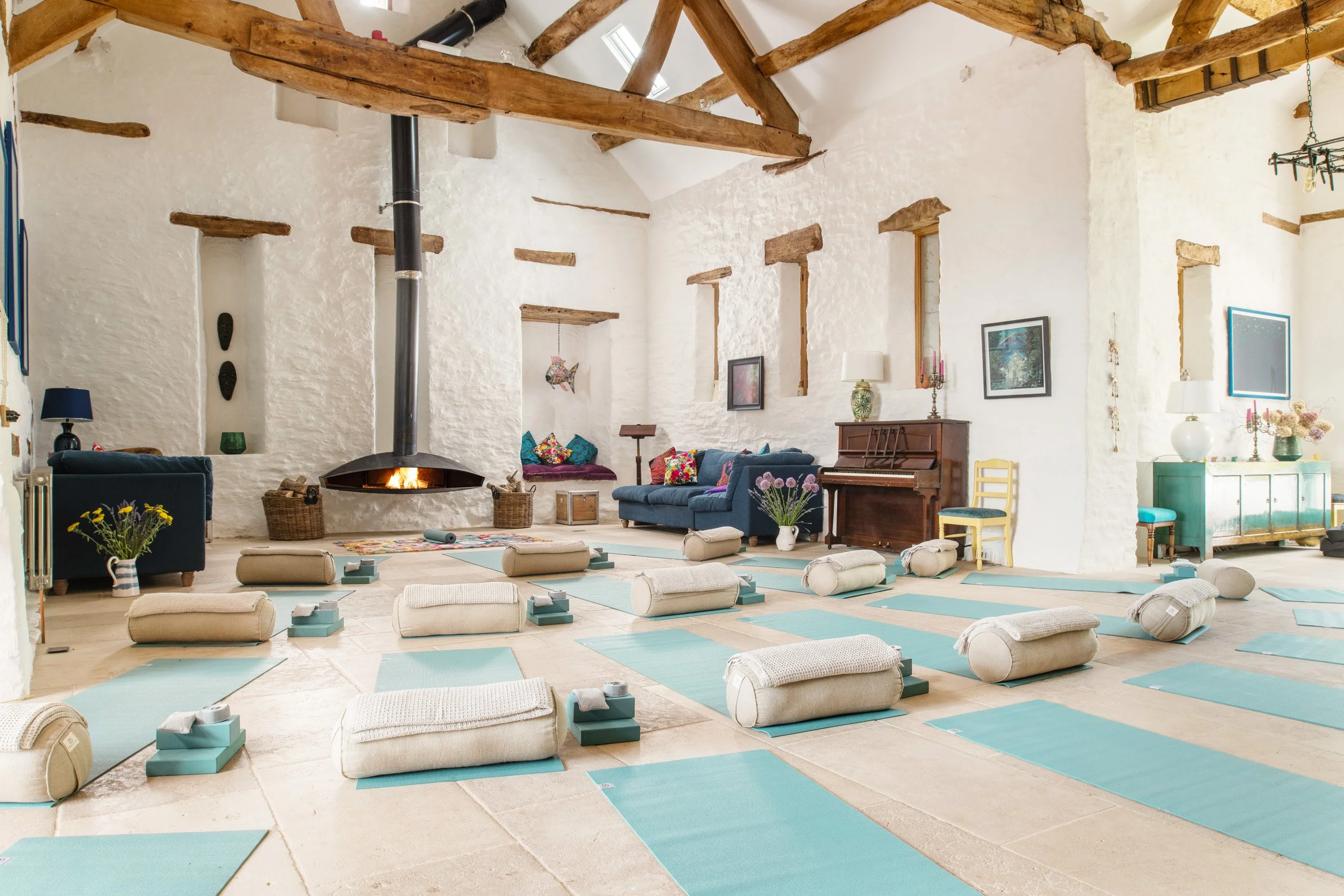 A spacious yoga studio decorated with white textured walls, exposed wooden beams, and large windows. The room is arranged with yoga mats, bolsters, and blocks on the floor, facing a modern black fireplace with a fire burning. There are colorful cushi