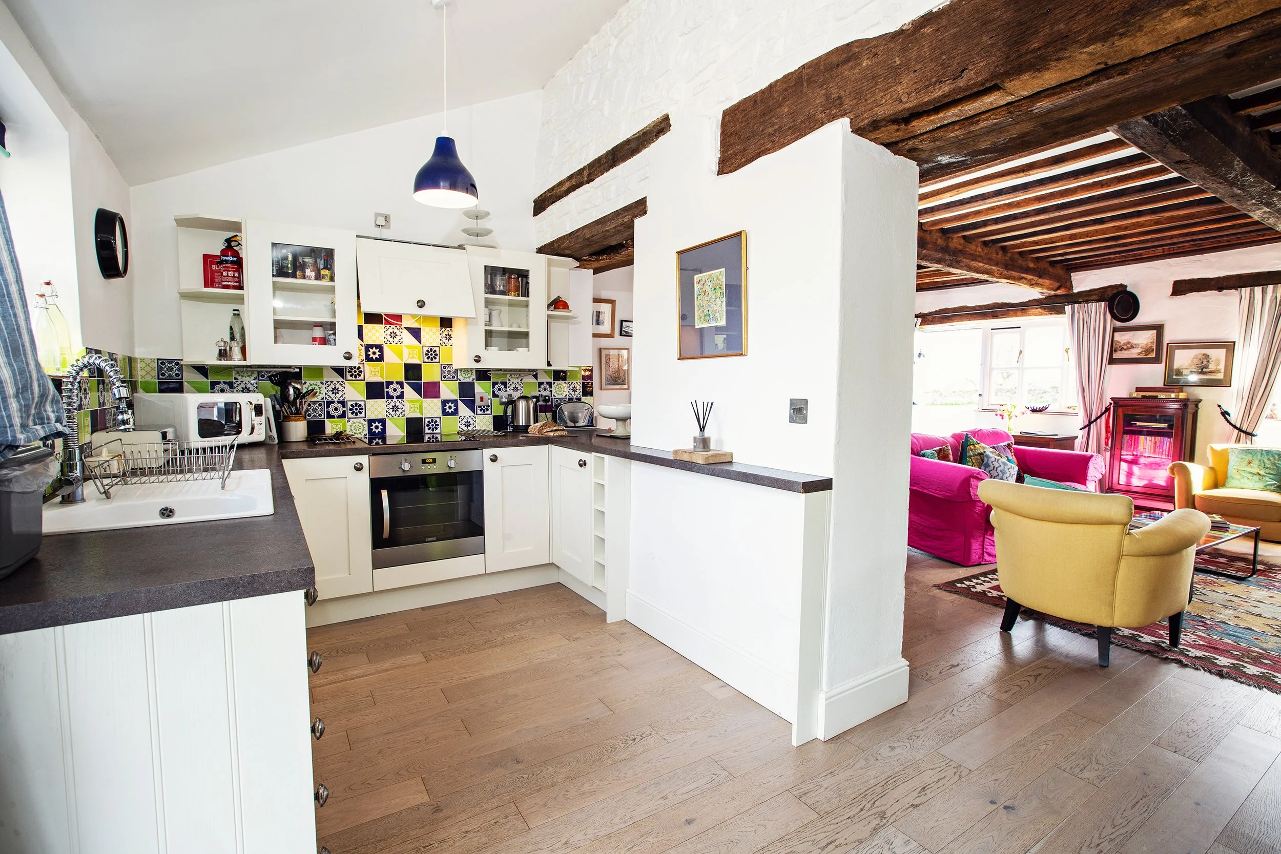 Open kitchen with white cabinets, a colorful tile backsplash, and a view into a living room with yellow and pink chairs, a sofa, and large windows.