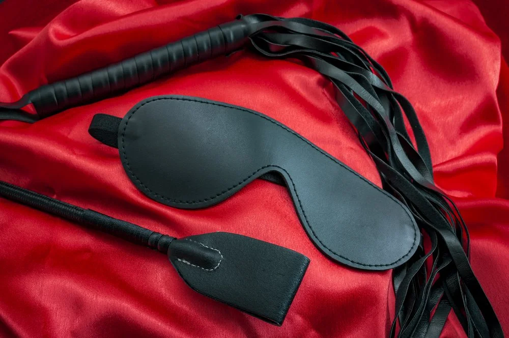 A black leather sleep mask, a black riding crop, and a black flogger with multiple tails on a red satin cloth.