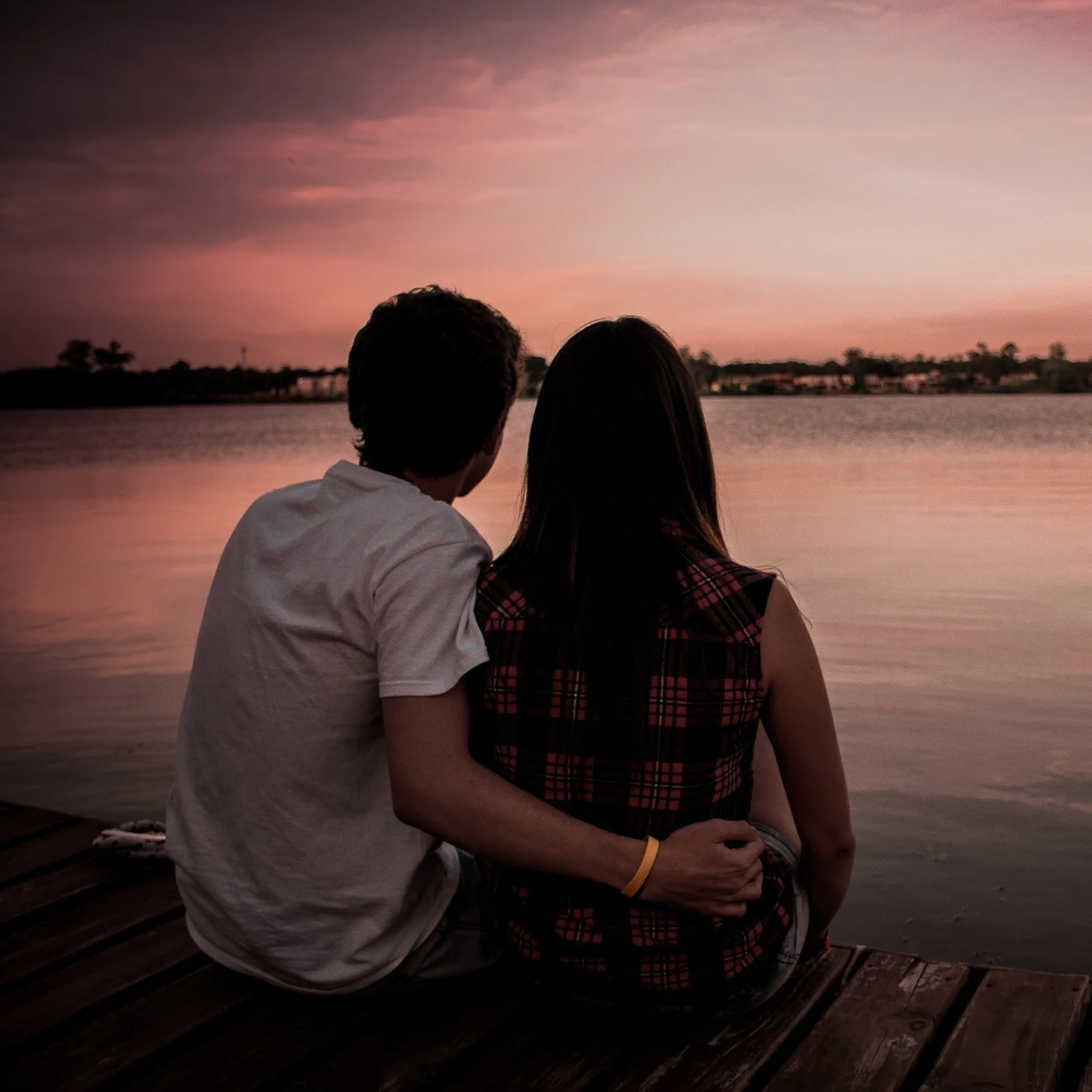A couple sitting on a wooden dock by the water at sunset, with a pink and purple sky reflected on the calm water.