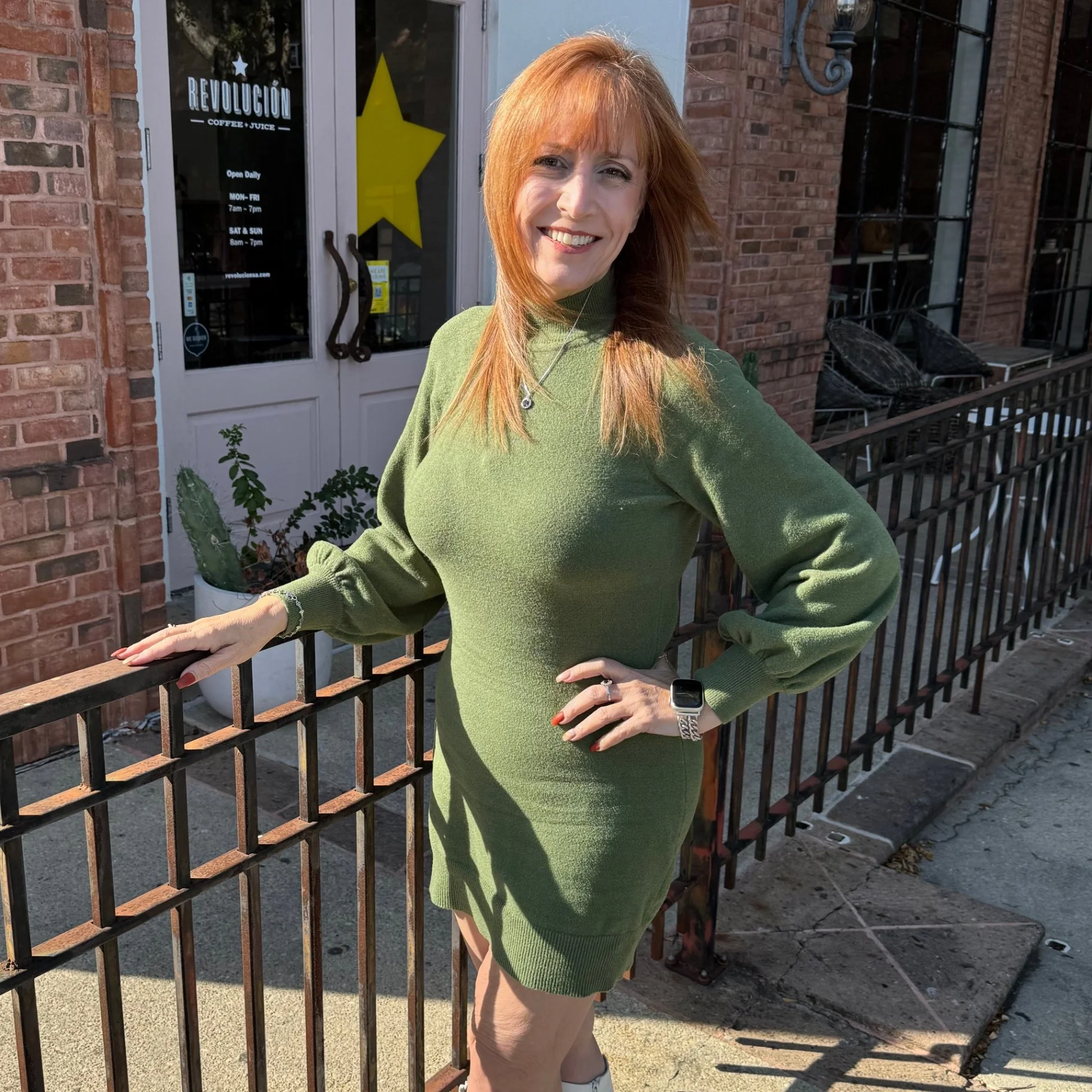 Woman with red hair wearing a green sweater dress, standing outdoors next to a black metal fence, smiling at the camera.