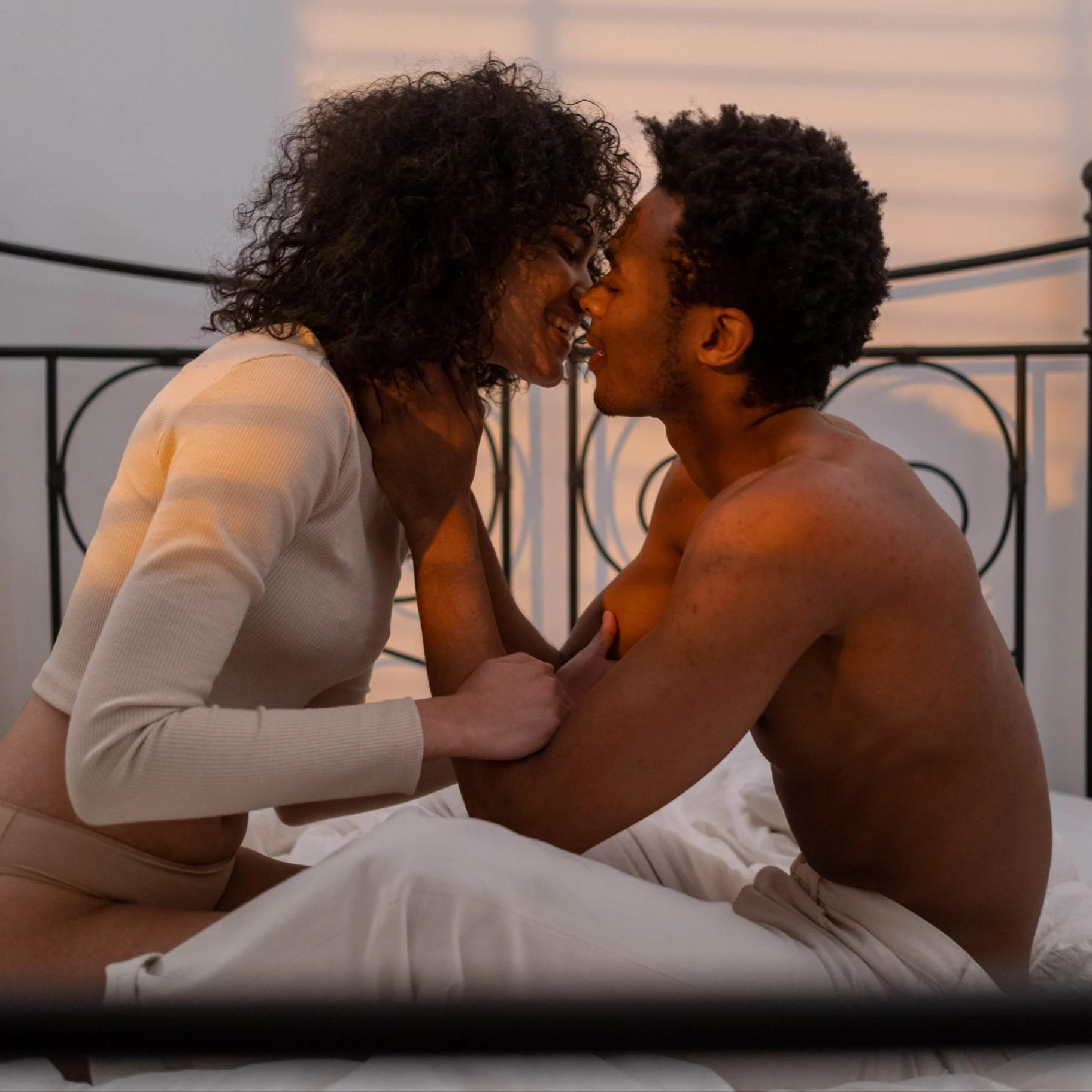A couple with dark skin and curly hair in a romantic moment on a bed, facing each other, with their faces close and eyes closed, holding each other's hands and touching each other's faces, at sunset with a metal headboard in the background.