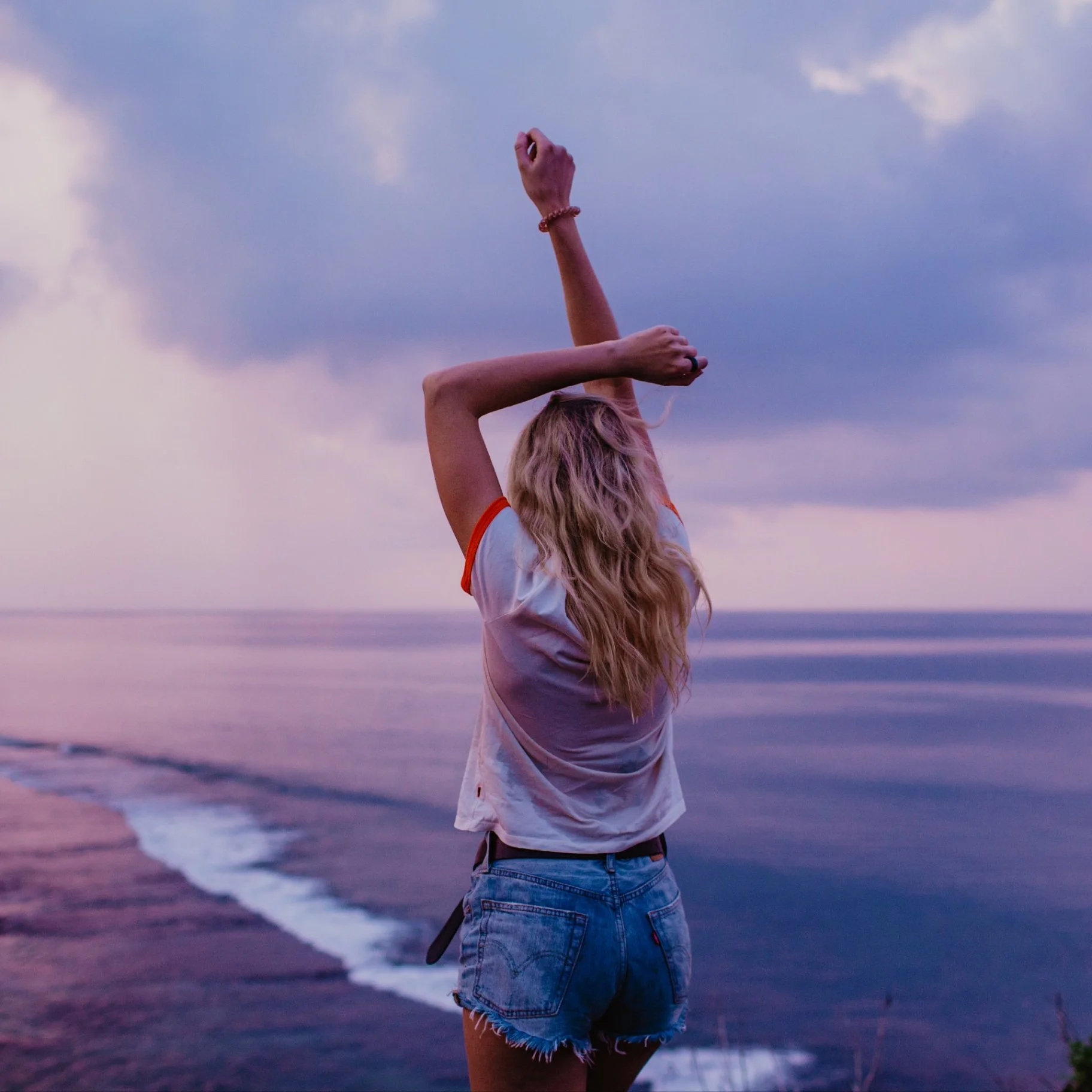 Woman standing by the ocean at sunset with arms raised, symbolizing healing and personal freedom