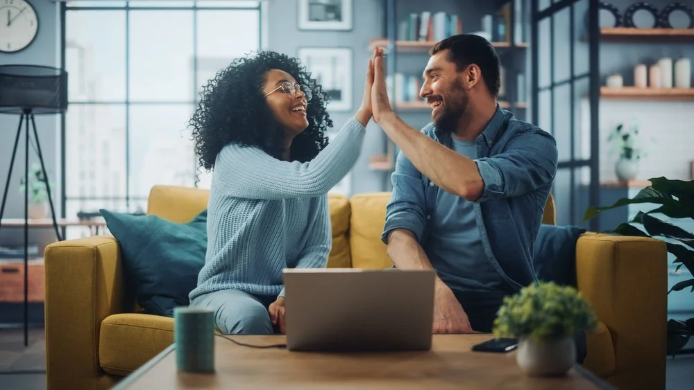 A man and woman are sitting on a yellow sofa, giving each other a high five and smiling in a modern, cozy living room.